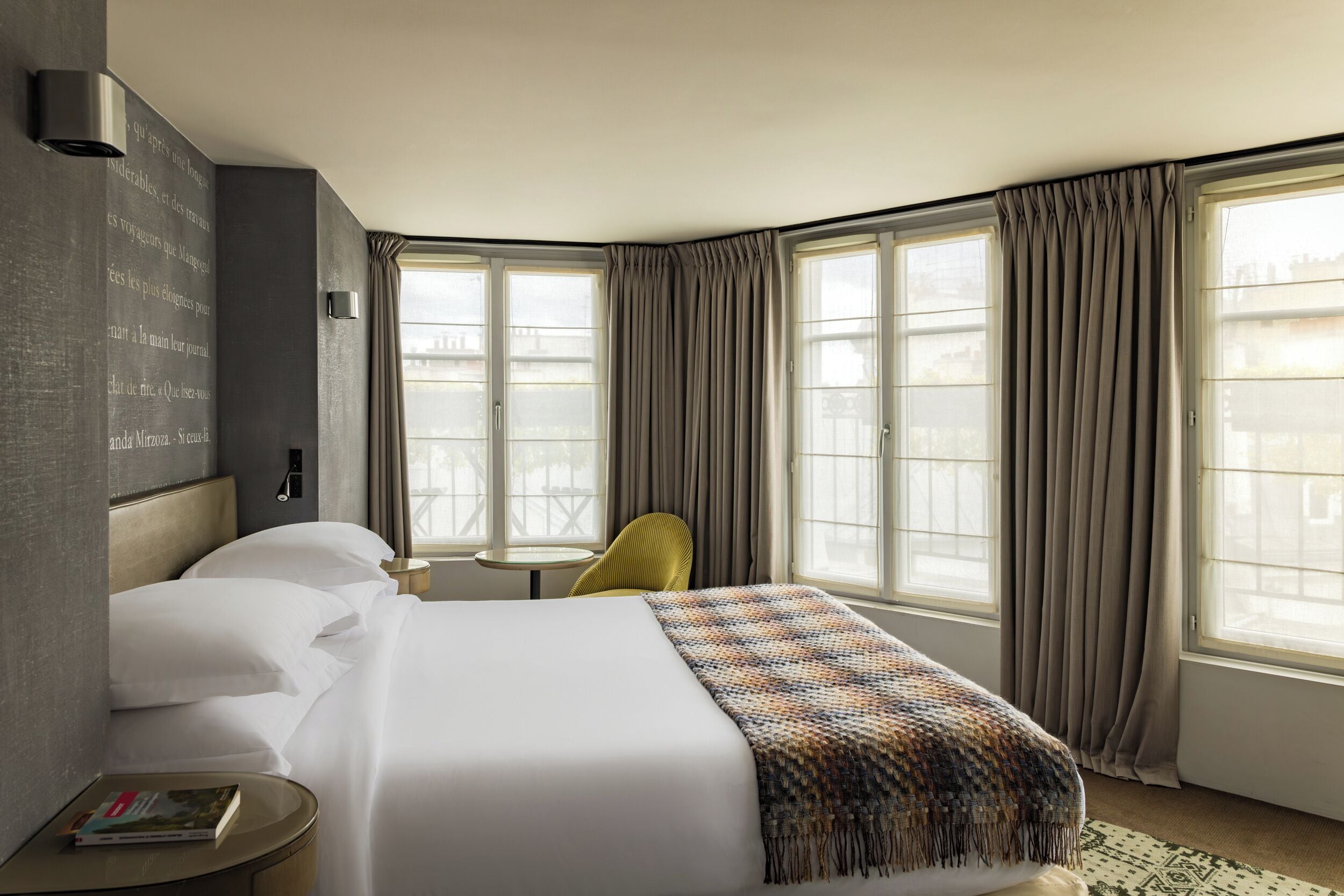 A hotel room with a large bed, a small round table, a chair, and a wall-mounted reading light.
