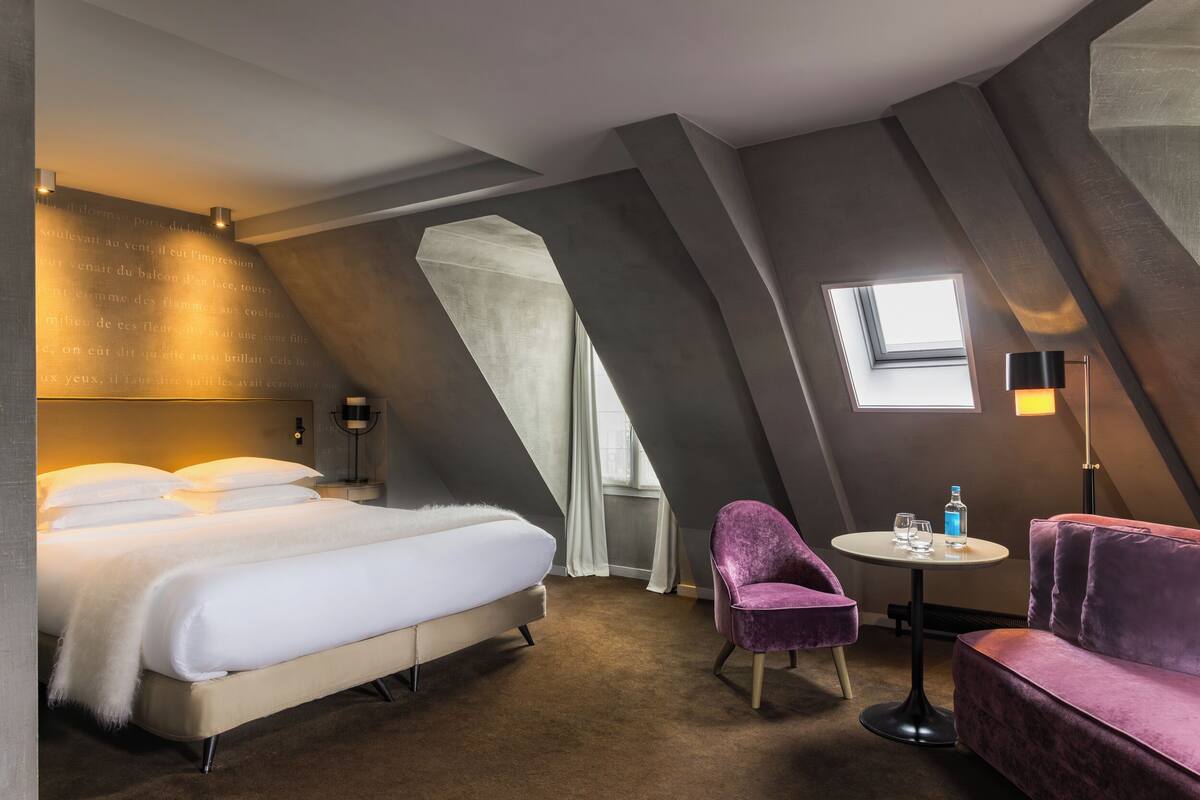Une chambre d’hôtel moderne avec un grand lit, une petite table ronde, un fauteuil violet et une fenêtre donnant sur le ciel.