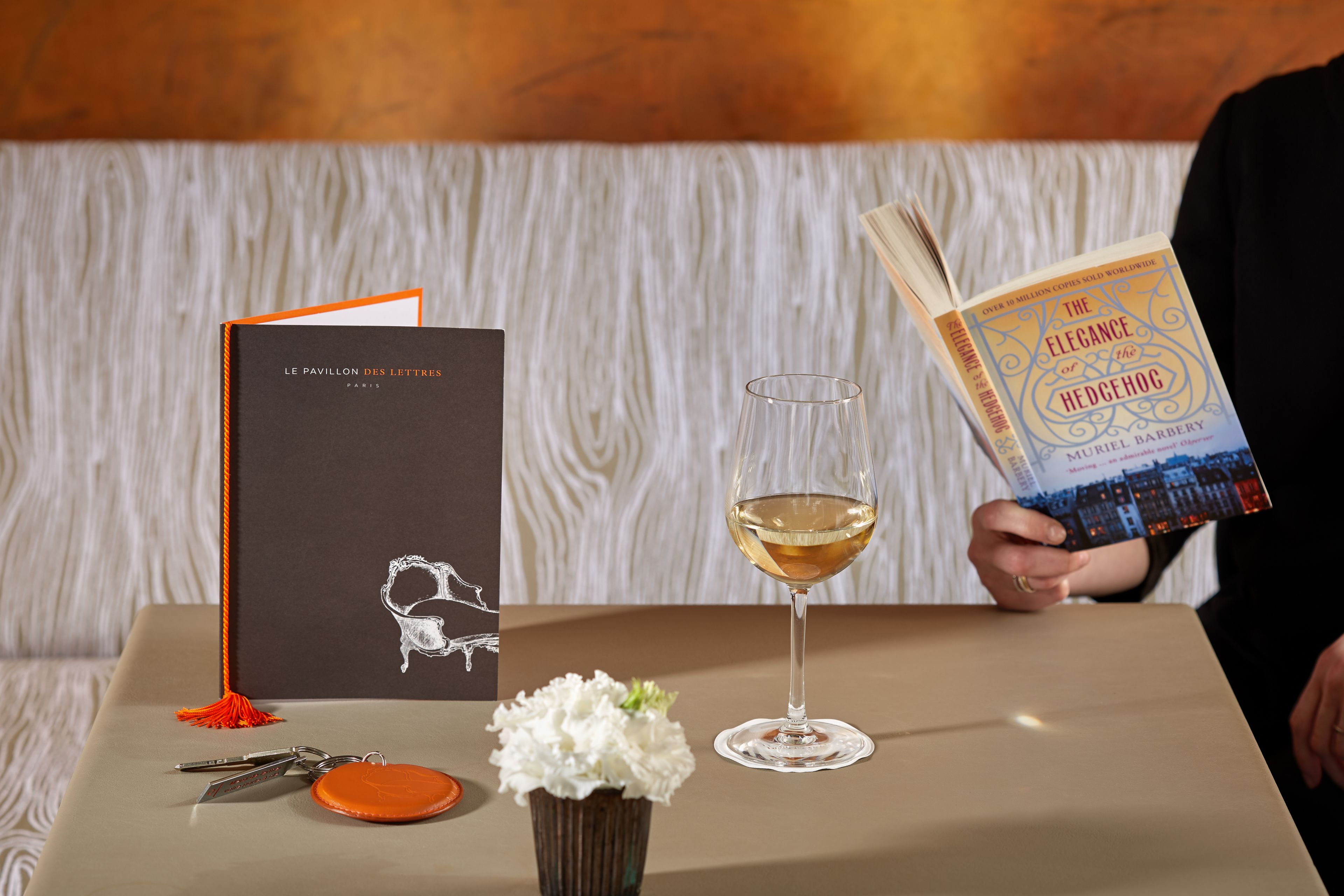 Ein Buch mit dem Titel „Die Eleganz des Igel“ auf einem Tisch zusammen mit einem Weinglas, einem Schlüsselanhänger und einer kleinen Vase mit Blumen.