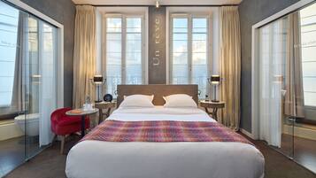 Quarto de hotel com uma cama grande, duas mesas de cabeceira, uma cadeira vermelha e uma janela ampla com cortinas.