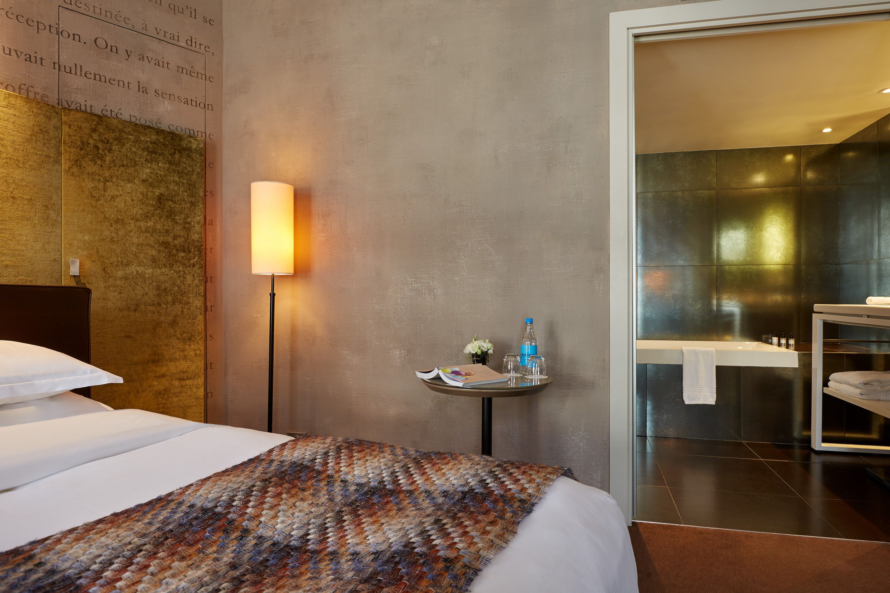 Ein Hotelzimmer mit einem ordentlich bezogenen Bett, einem Nachttisch mit Lampe, einem kleinen Tisch mit Buch und einer Blumenvase, sowie einem durch eine geöffnete Tür einsehbaren Badezimmer.