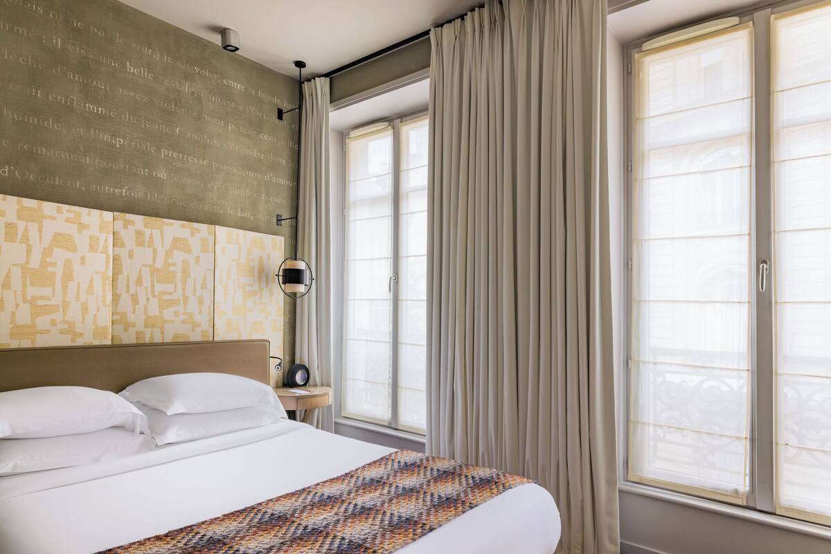 Une chambre d’hôtel avec un grand lit, une tête de lit, une table de chevet avec une lampe et une fenêtre avec des rideaux.