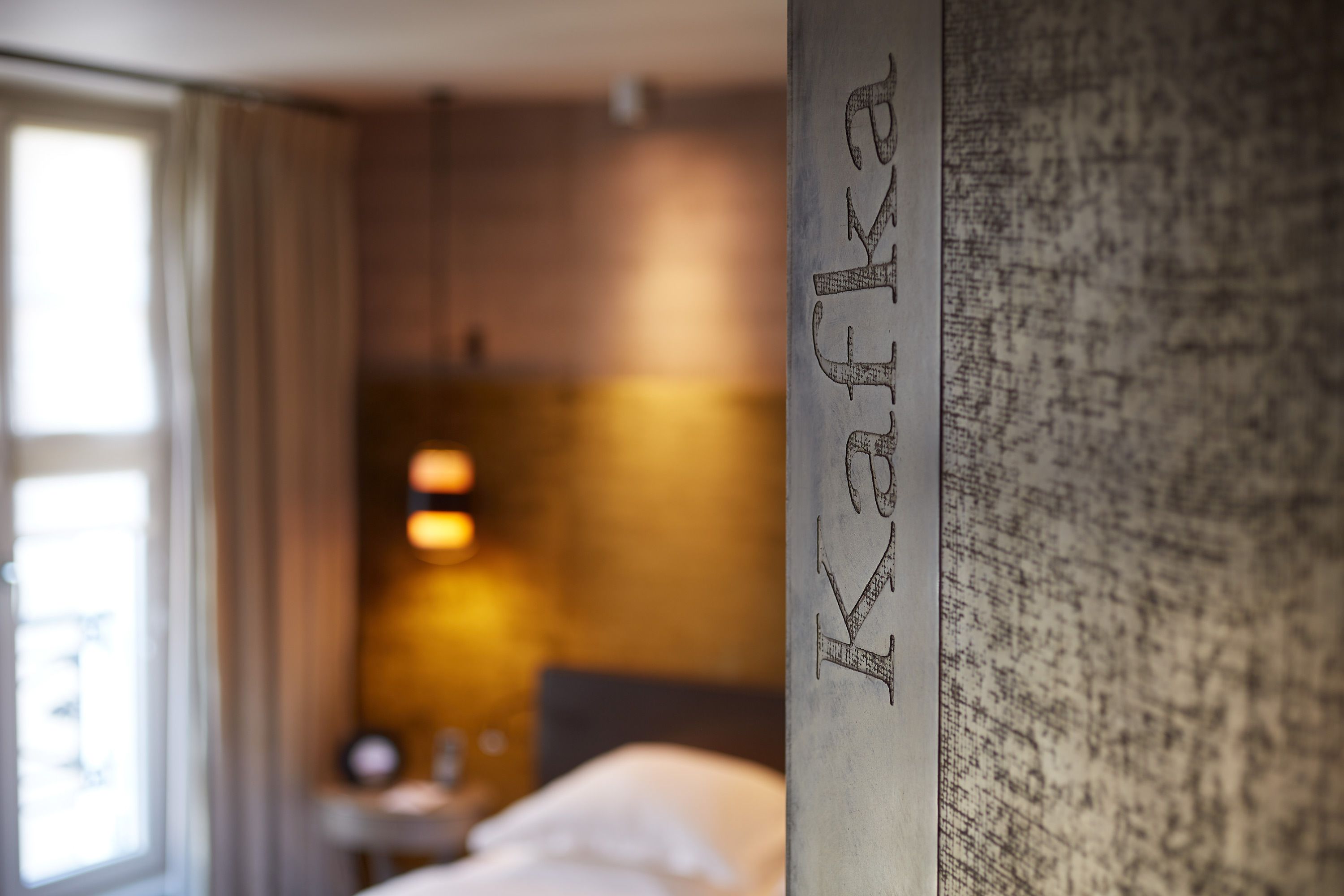 Una habitación de hotel con cama, mesita de noche y lámpara. La habitación tiene una ventana con cortinas y una pared donde se lee la palabra "Kafka".