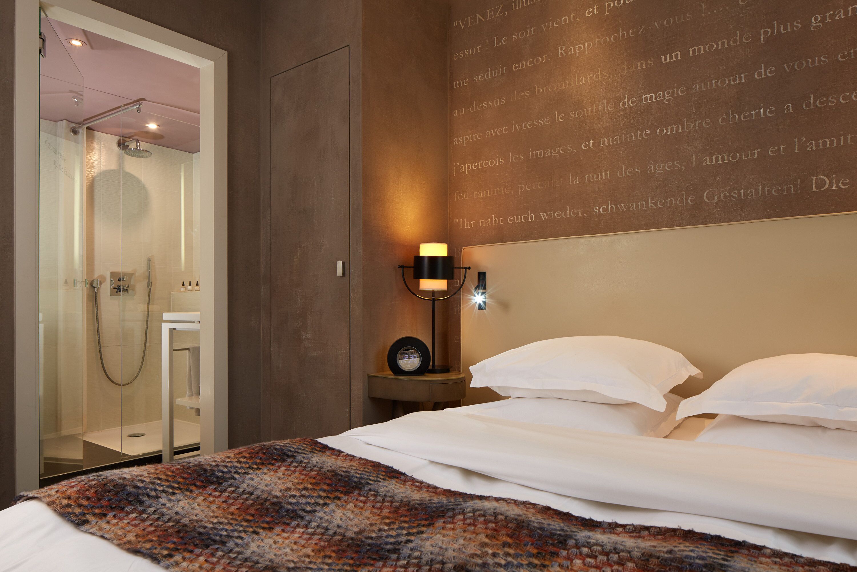 A hotel room with a glass-enclosed shower, a bedside table with a lamp, and a bed with white bedding.
