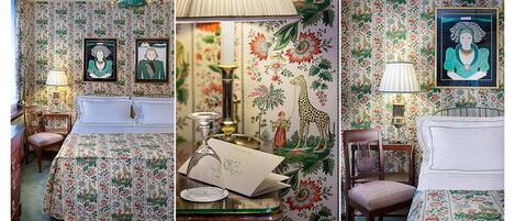 A room with floral wallpaper, a bed with a matching headboard, a desk with a lamp, and framed artwork on the wall.