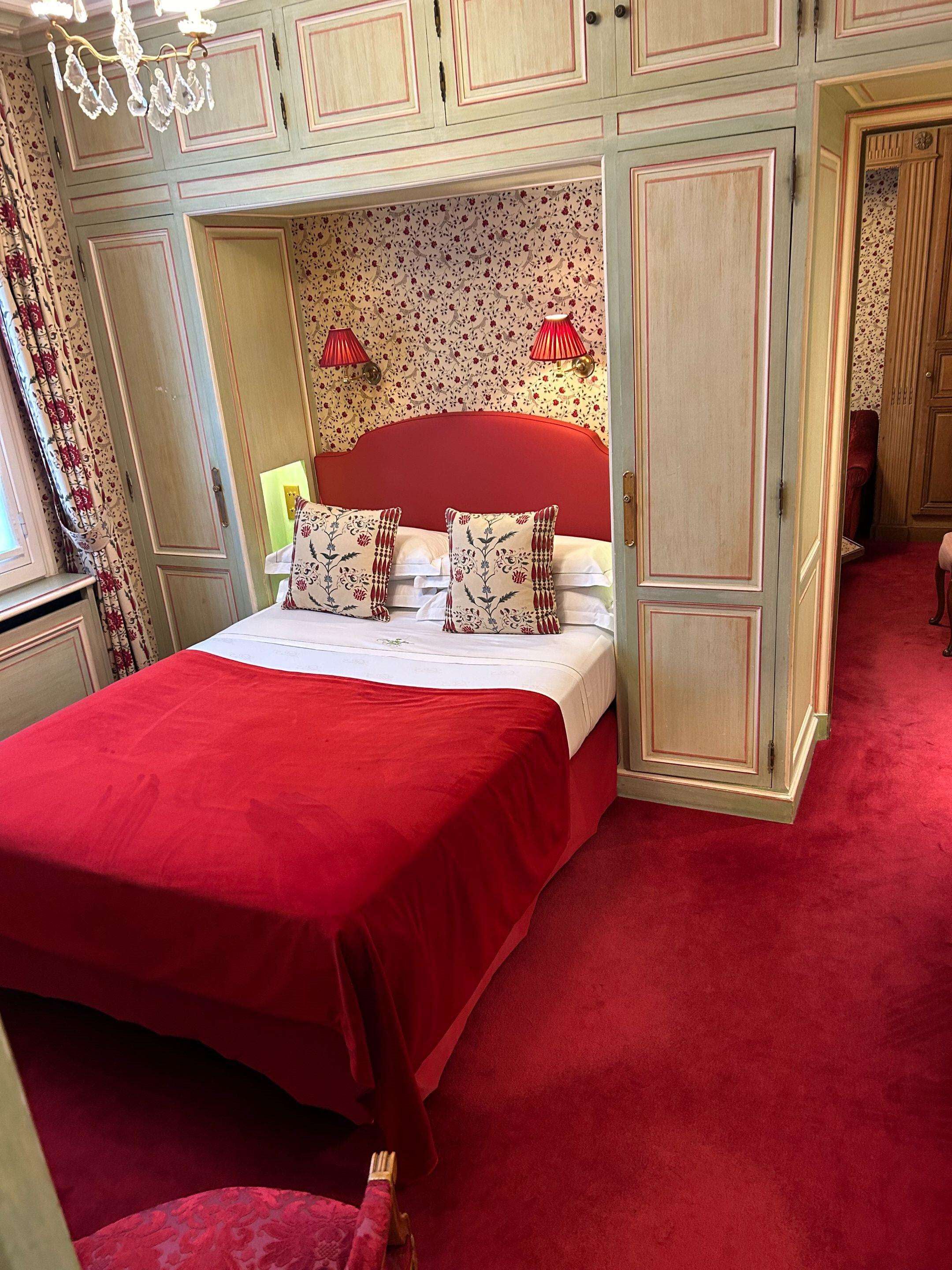A bedroom with a red bedspread, floral patterned pillows, and a red headboard. There is a chandelier above the bed, and the room has a red carpet.
