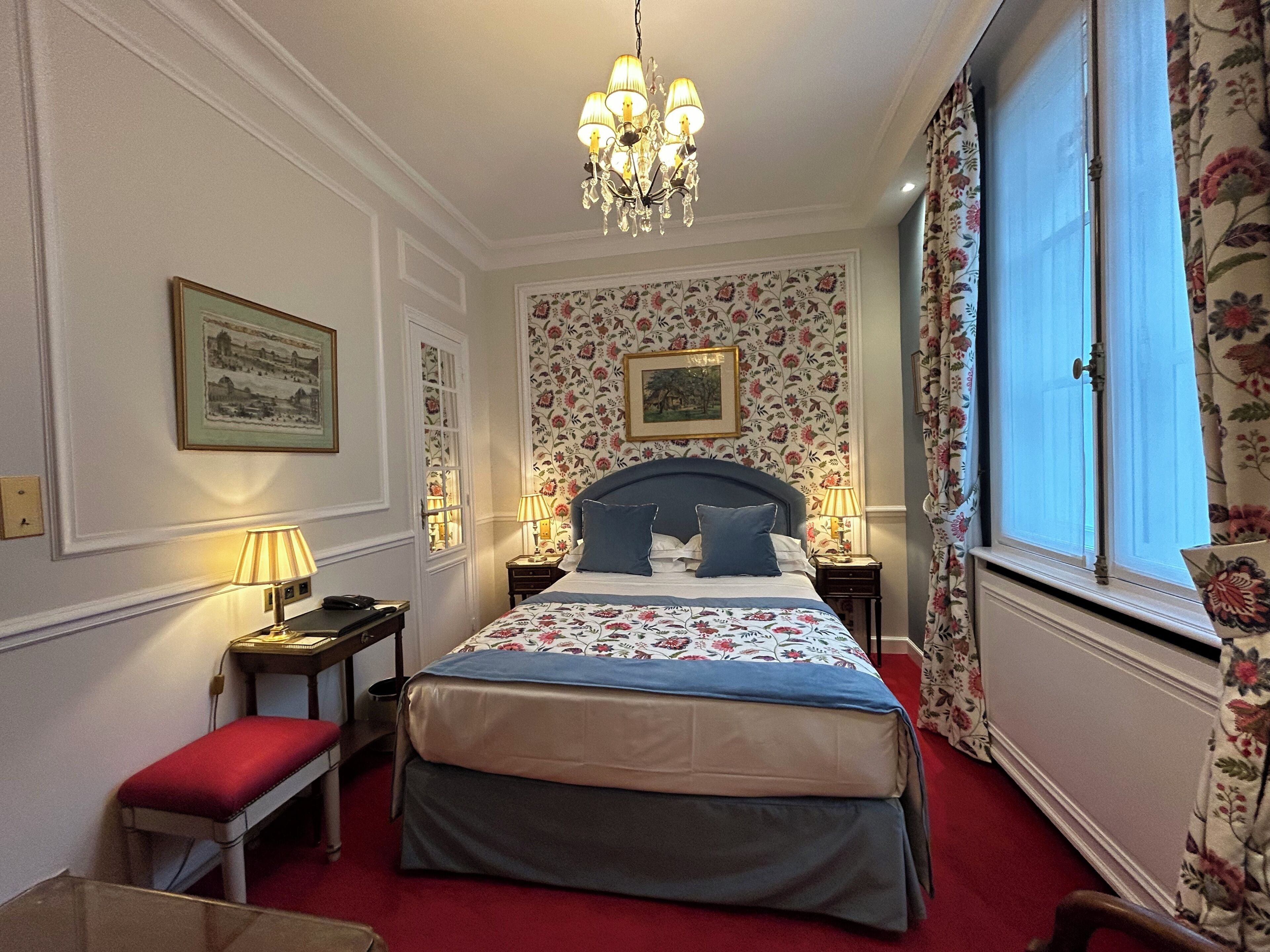 Un dormitorio con cama, un banco, un escritorio, una silla, una araña y una ventana con cortinas.