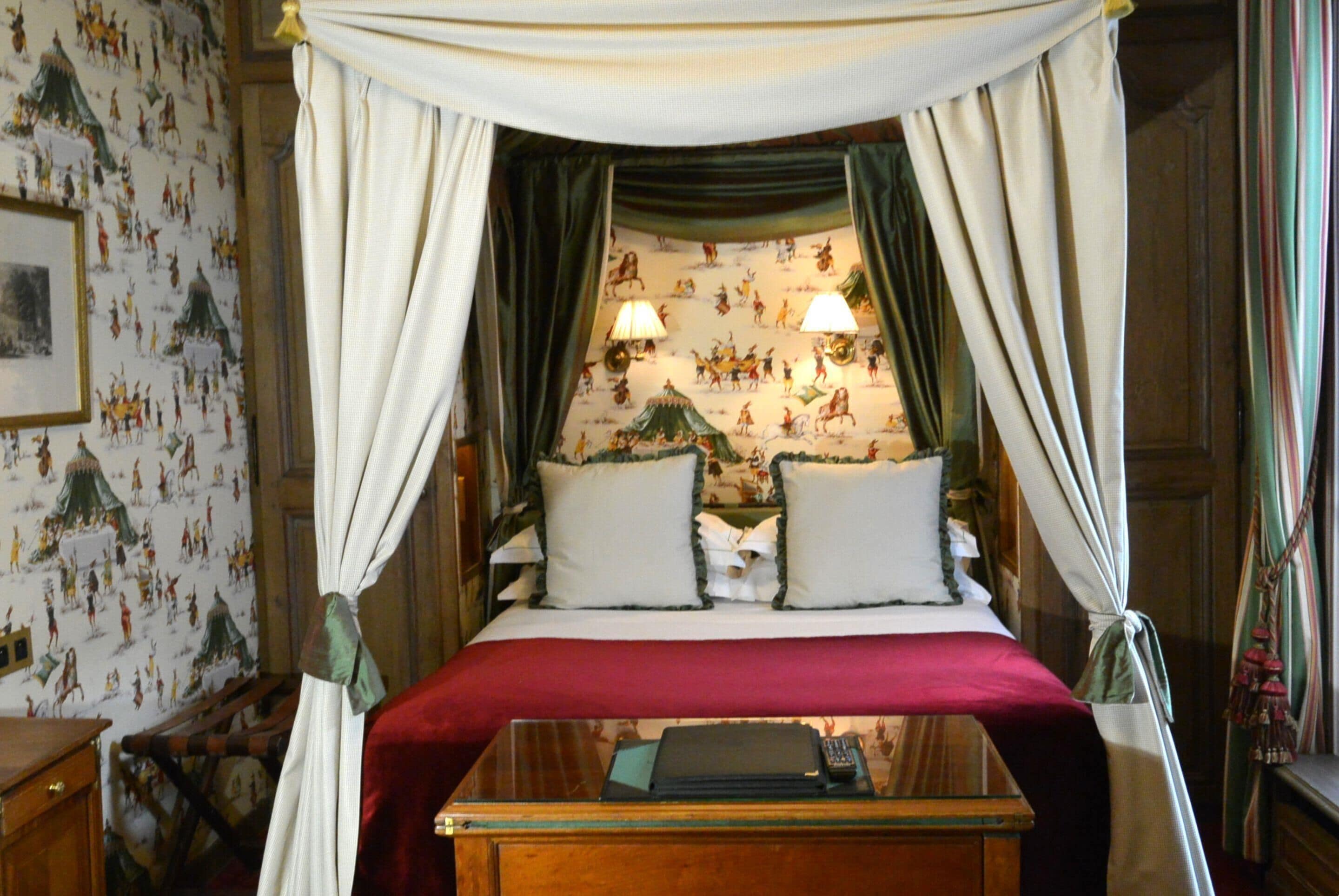 A four-poster canopy bed with a red bedspread, two pillows, and a bedside table with a lamp. The room has patterned wallpaper and a wooden nightstand.