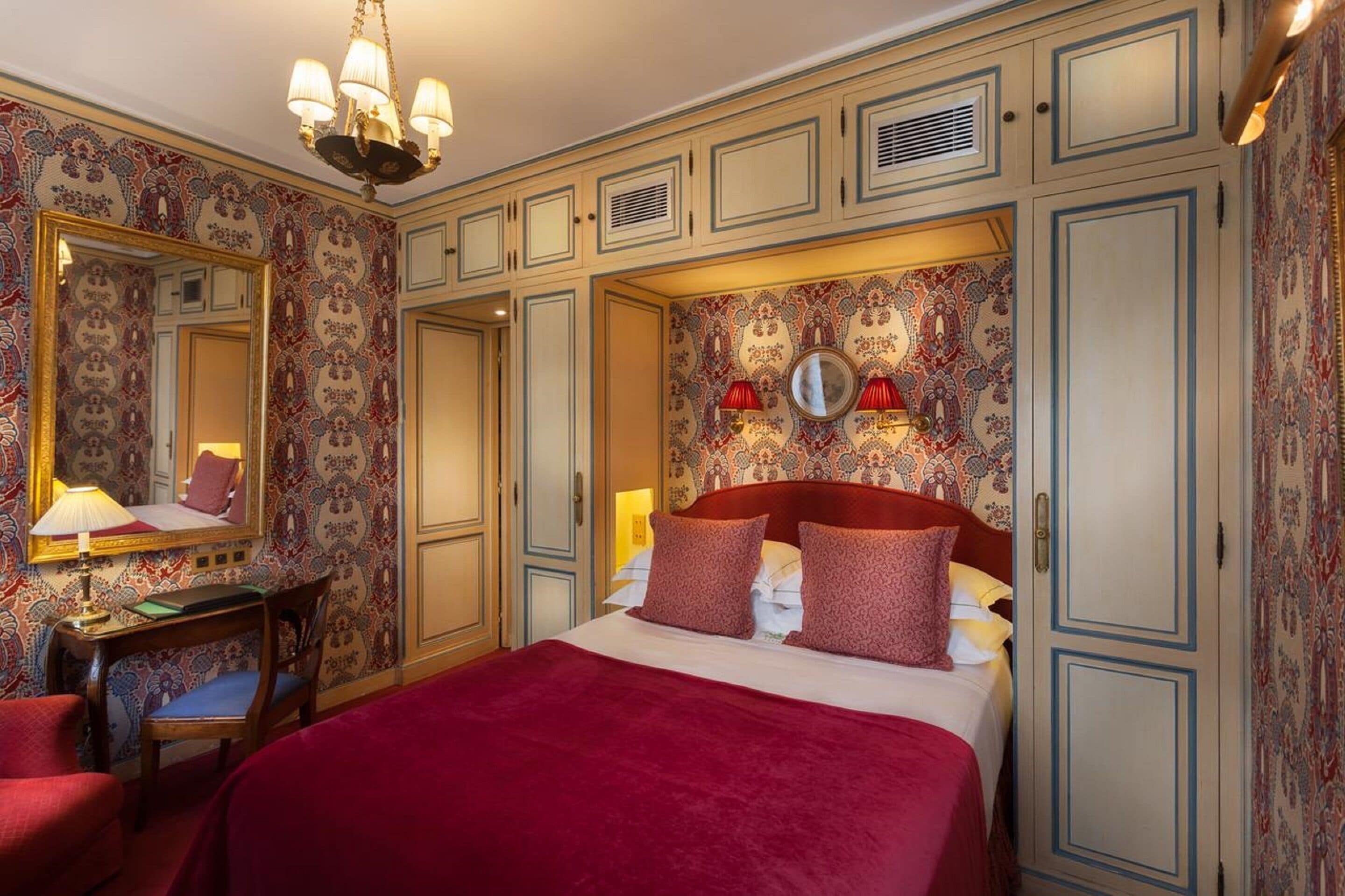 A hotel room with a large bed, a desk with a chair, a mirror, and a patterned wallpaper.