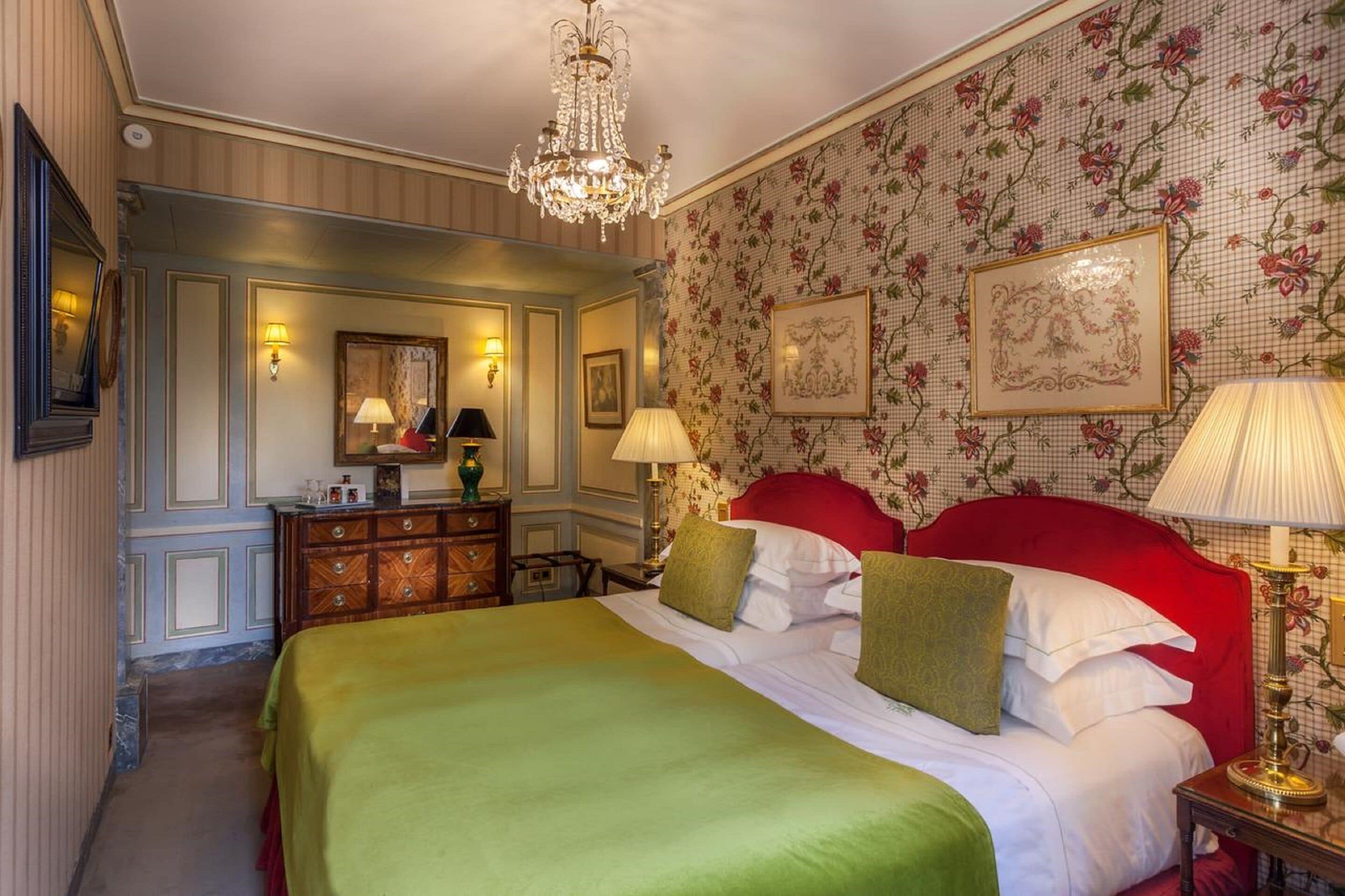 Ein Hotelzimmer mit einem großen Bett, einer Kristallleuchter, blumigem Wandpapier und einem roten Kopfteil.