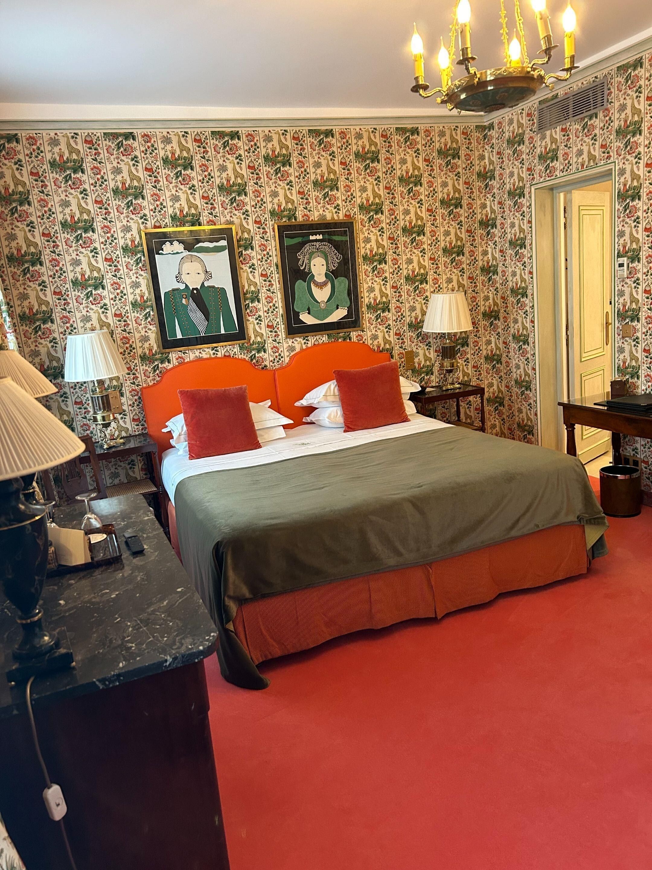 A hotel room with a red carpet, a bed with orange and gray bedding, a wooden headboard, and a patterned wallpaper.