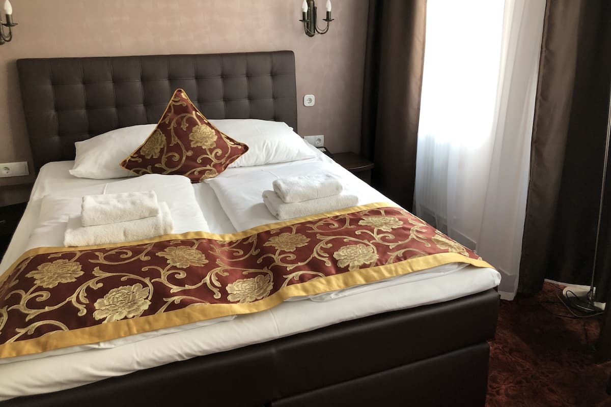 A neatly made bed with a dark headboard, white and gold patterned bedding, and a red and gold throw blanket.