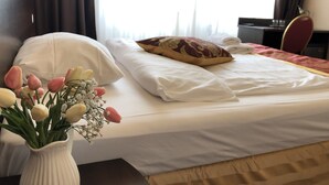 A neatly made bed with white linens and a decorative pillow, a vase of flowers on a bedside table, and a window with curtains. - Hotel Atlas Berlin (Berlin)