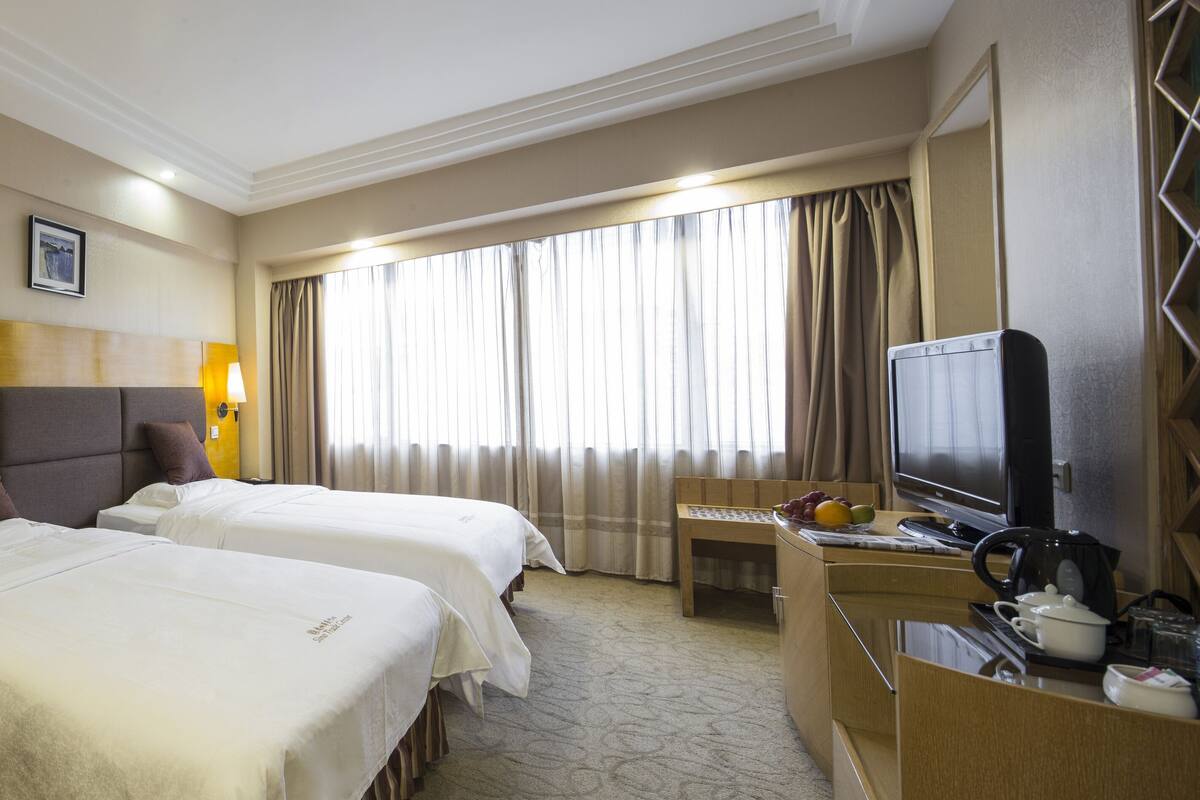 A hotel room with two beds, a TV, a desk with a chair, and a window with curtains.