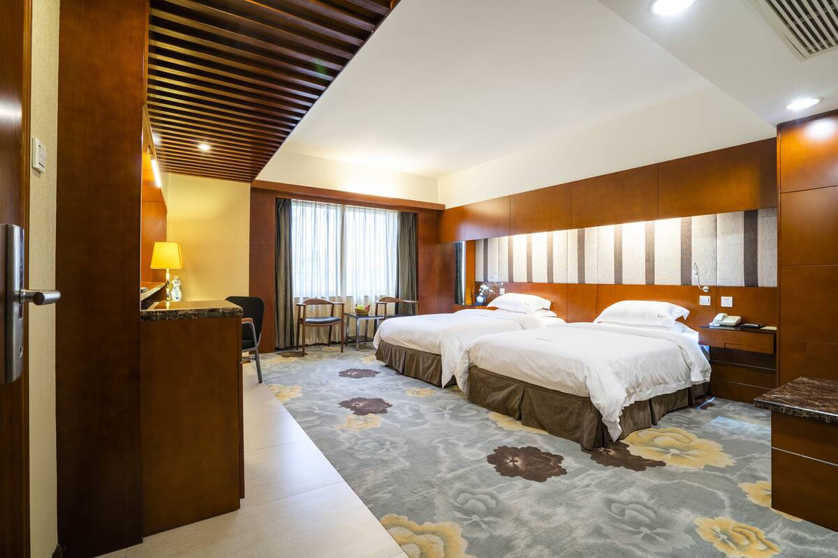 A hotel room with two beds, a desk with a lamp, a chair, and a large window with curtains.