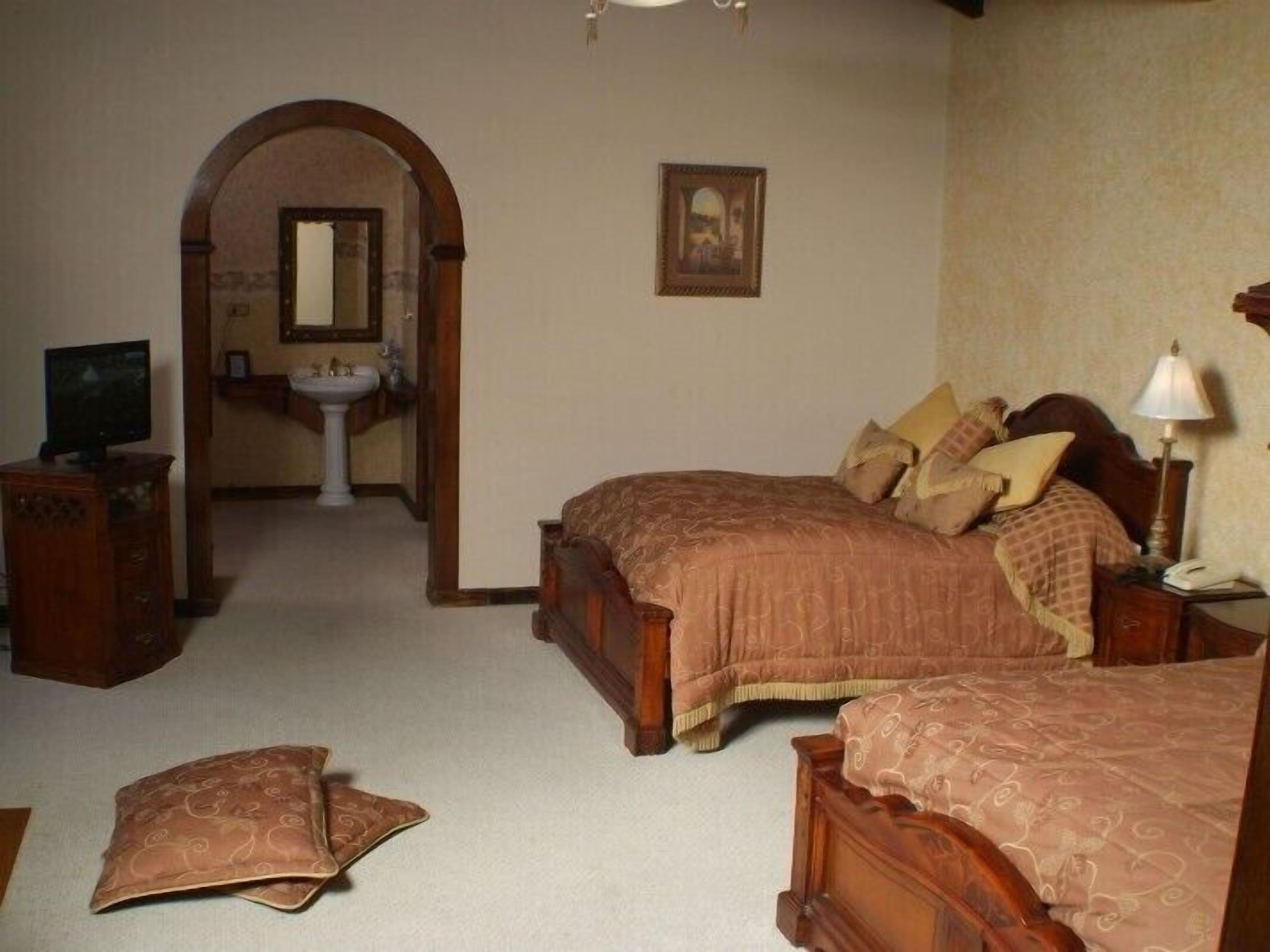A bedroom with two beds, a wooden dresser, a television, a mirror, and a bathroom visible through an archway.