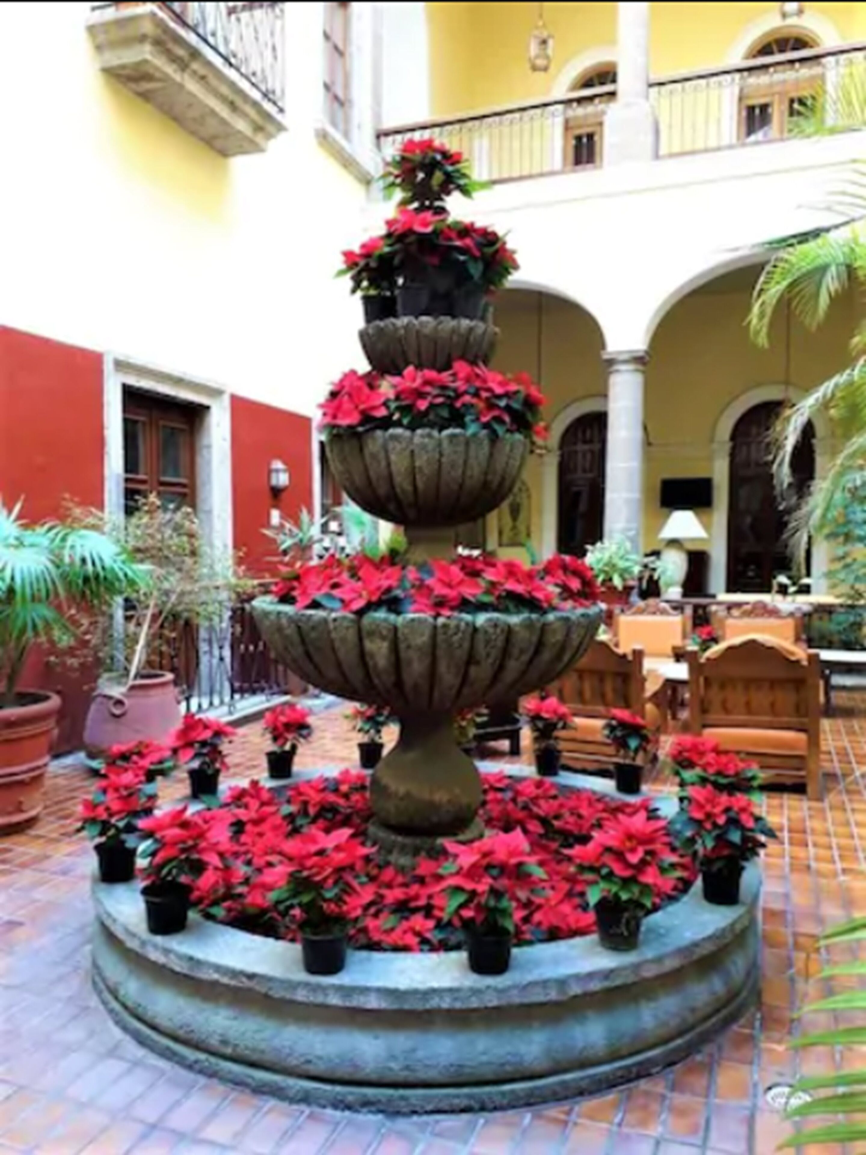 A multi-tiered stone fountain with red flowers in a courtyard with tiled flooring, surrounded by potted plants and outdoor furniture.
