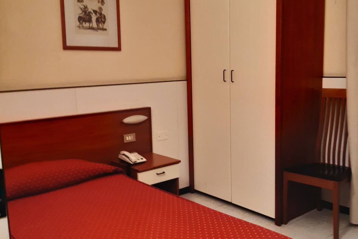Un dormitorio con cama, armario, silla, mesita de noche con lámpara y un cuadro en la pared.