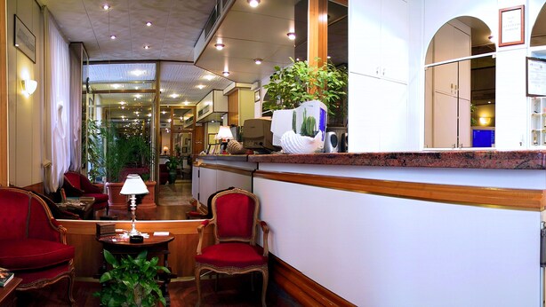 A hotel lobby with a reception desk, red upholstered chairs, a coffee table, and a potted plant. - Hotel Mayorca (Milan)