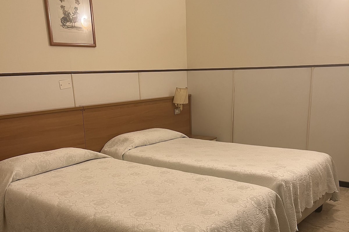 Habitación de hotel con dos camas individuales, un banco de madera, una lámpara de pared y un cuadro enmarcado.