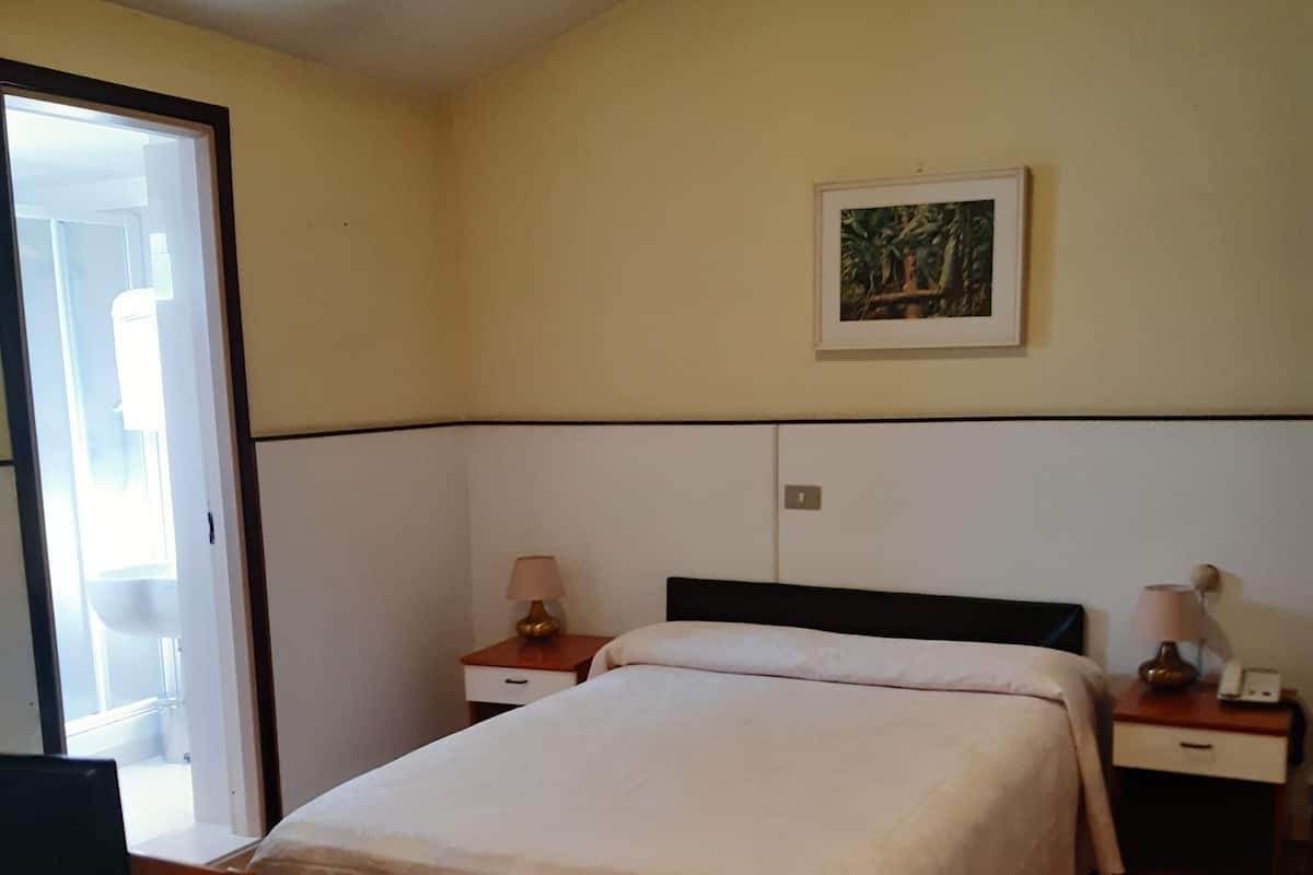 Un dormitorio con una cama, dos mesitas de noche, una lámpara, un cuadro enmarcado en la pared y una puerta que da a otra habitación.