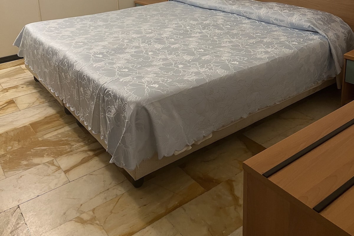 Un dormitorio con cama, mesita de noche, banco de madera, cuadro en la pared y suelo de baldosas.