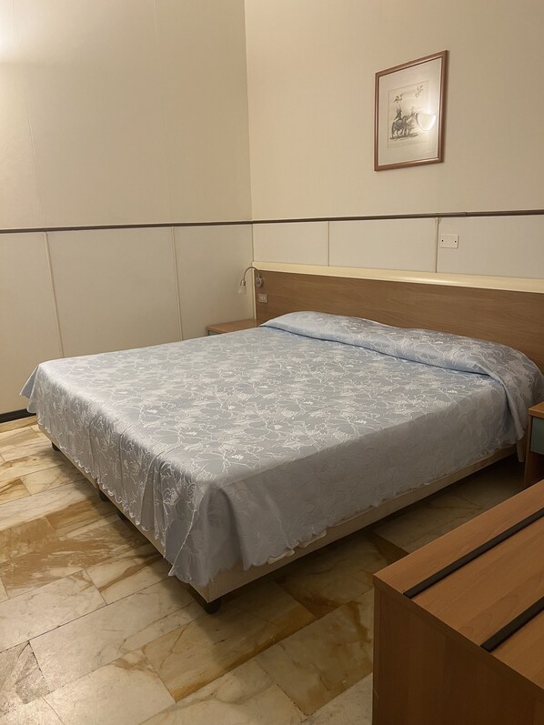 A bedroom with a bed, a nightstand, a wooden bench, a wall-mounted picture, and a tiled floor.