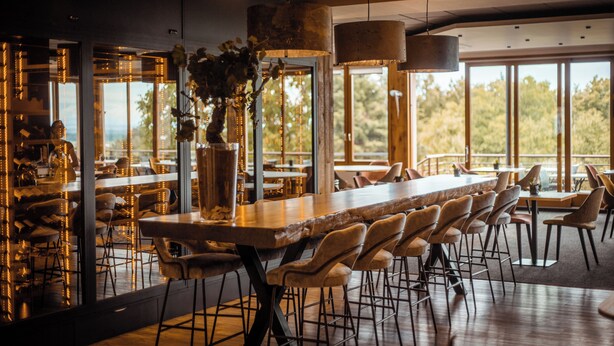 A restaurant with a long wooden bar, multiple bar stools, and large windows offering a view of the outdoors. - Hotel du Bollenberg (Westhalten)