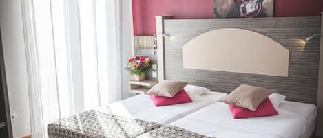 A hotel room with a bed, a bedside table, a nightstand, a window with sheer curtains, and a painting on the wall.
