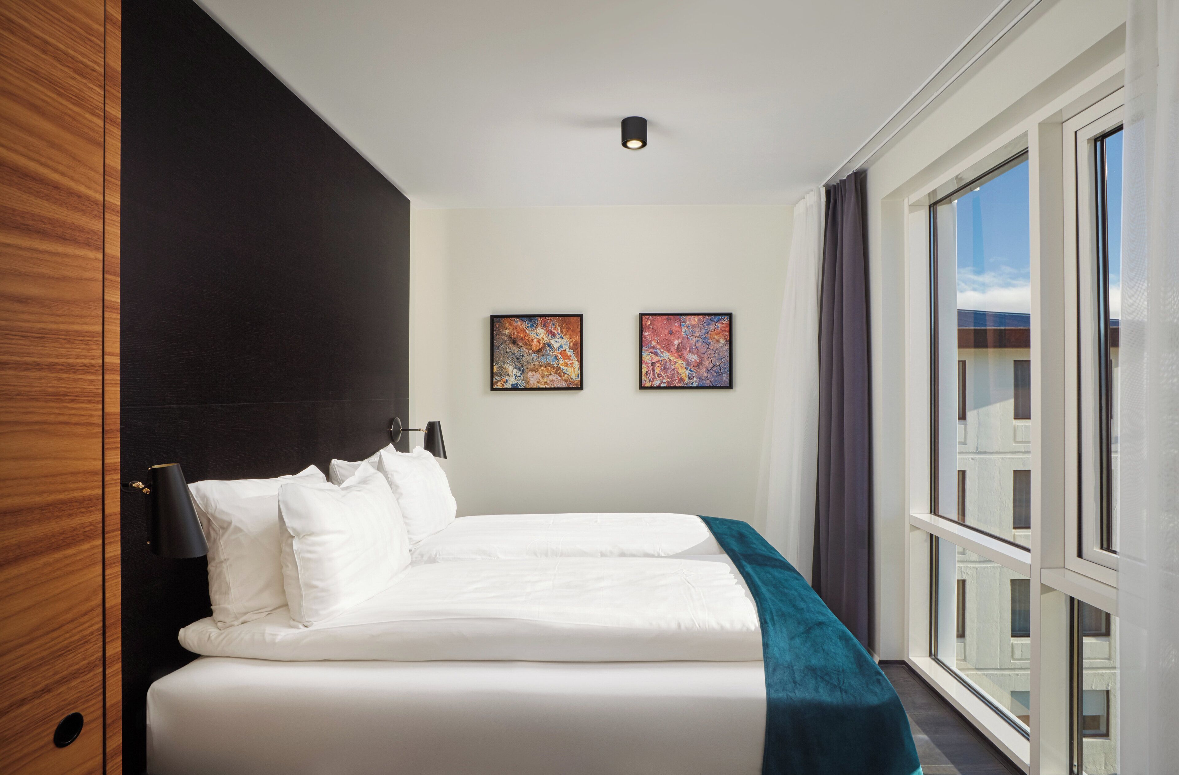 A hotel room with a large bed, two framed artworks on the wall, and a view of the city through the window.