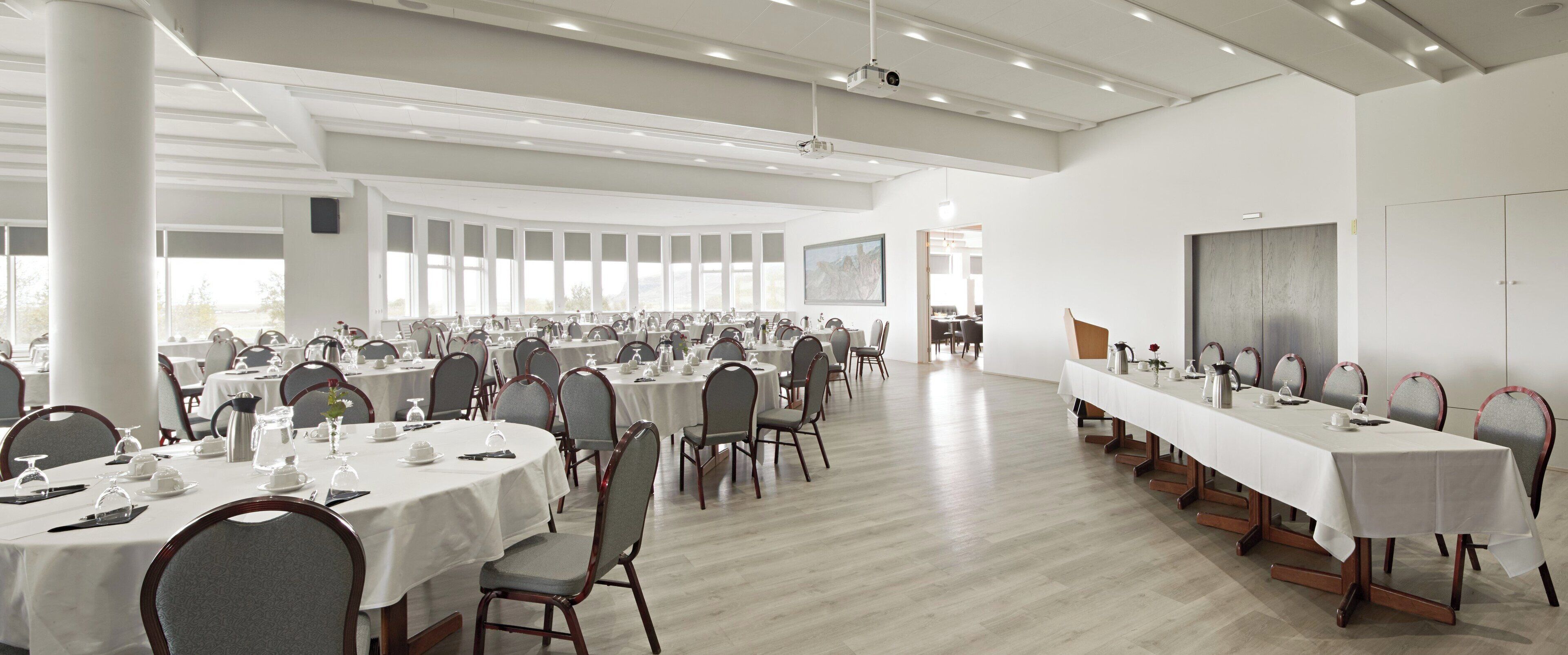 A large conference room with multiple round tables, each set with white tablecloths and chairs, and a long table with a white tablecloth and chairs.