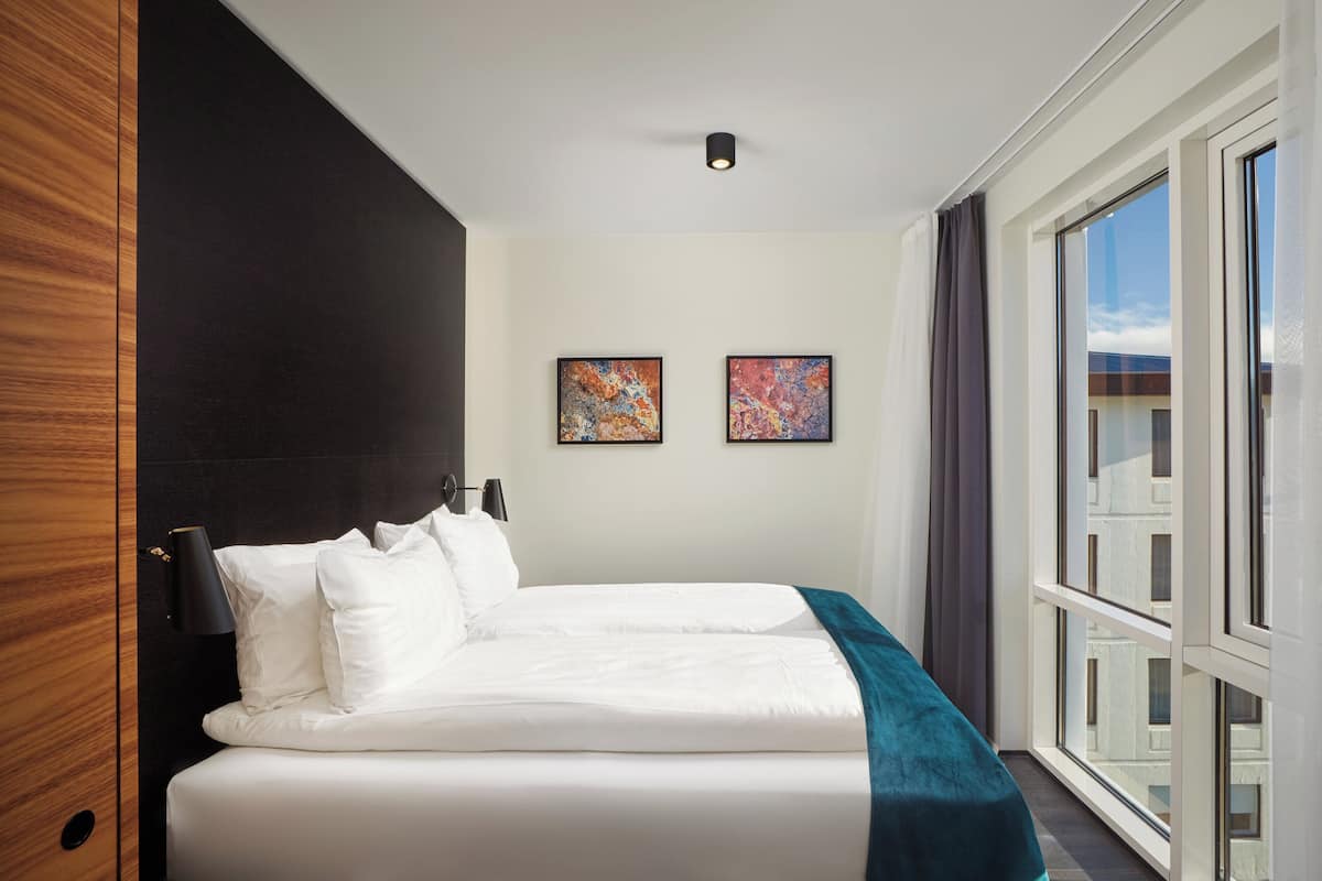 A hotel room with a large bed, two framed artworks on the wall, and a view of the city through the window.