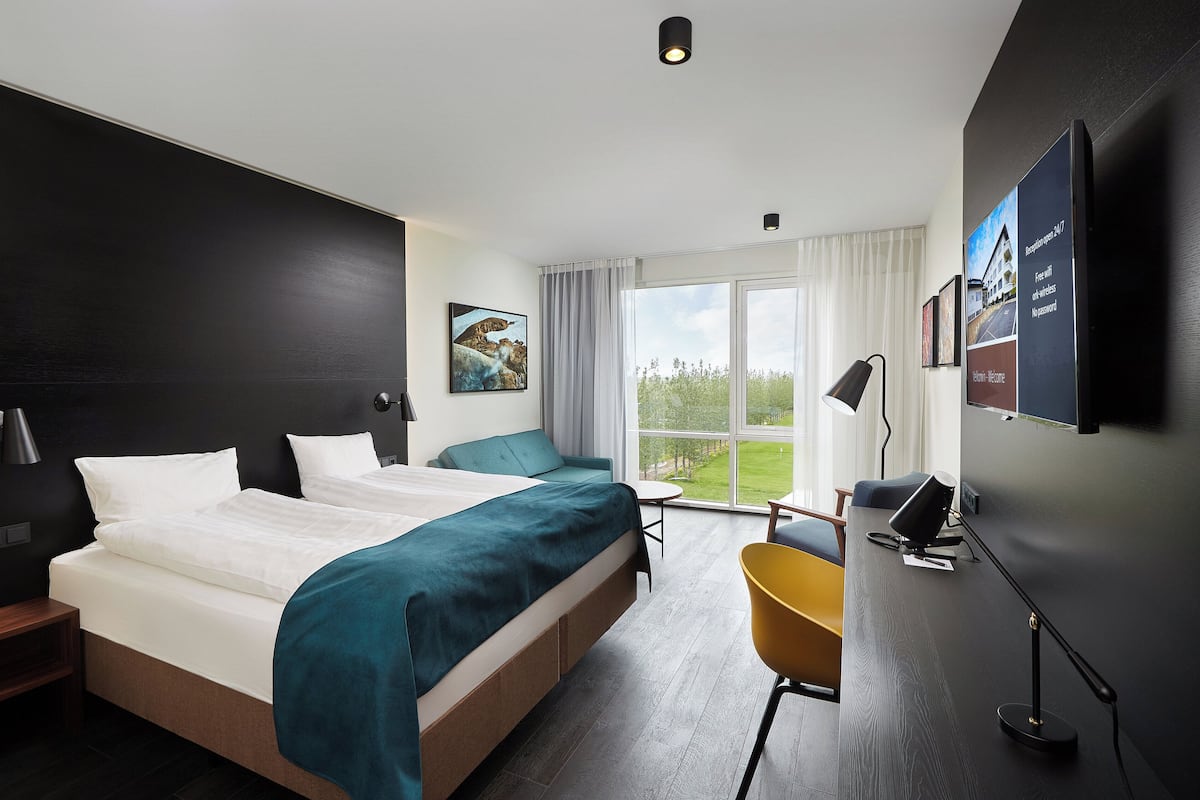 A modern hotel room with a large bed, a desk, a chair, and a large window offering a view of the outdoors.