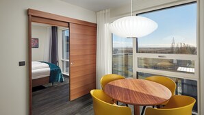 A modern hotel room with a large window, a wooden door, a bed with a teal blanket, a round wooden table, and yellow chairs. - Hotel Örk (Hveragerdi)