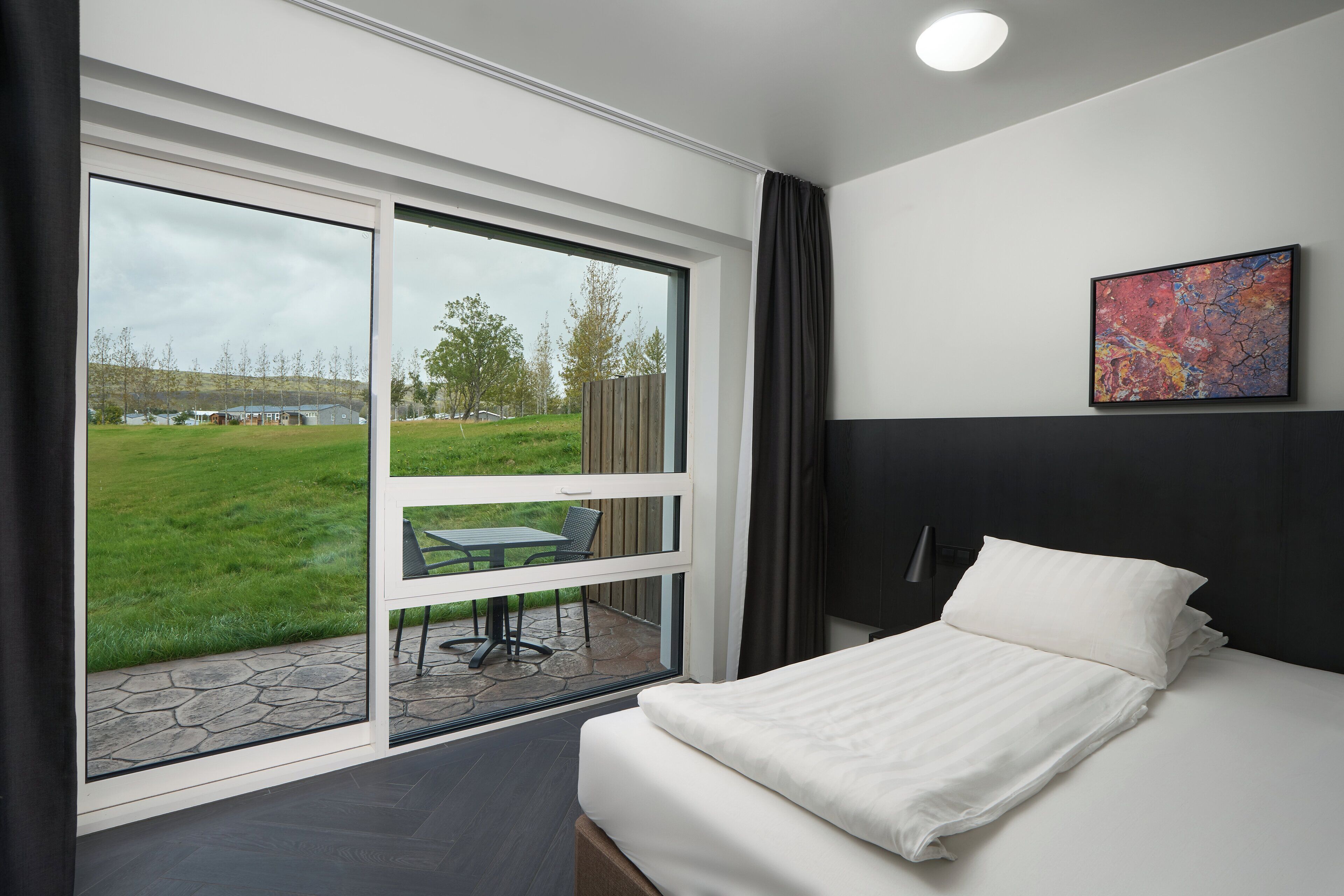 A modern hotel room with a large sliding glass door leading to an outdoor patio, a neatly made bed, and a single abstract painting on the wall.