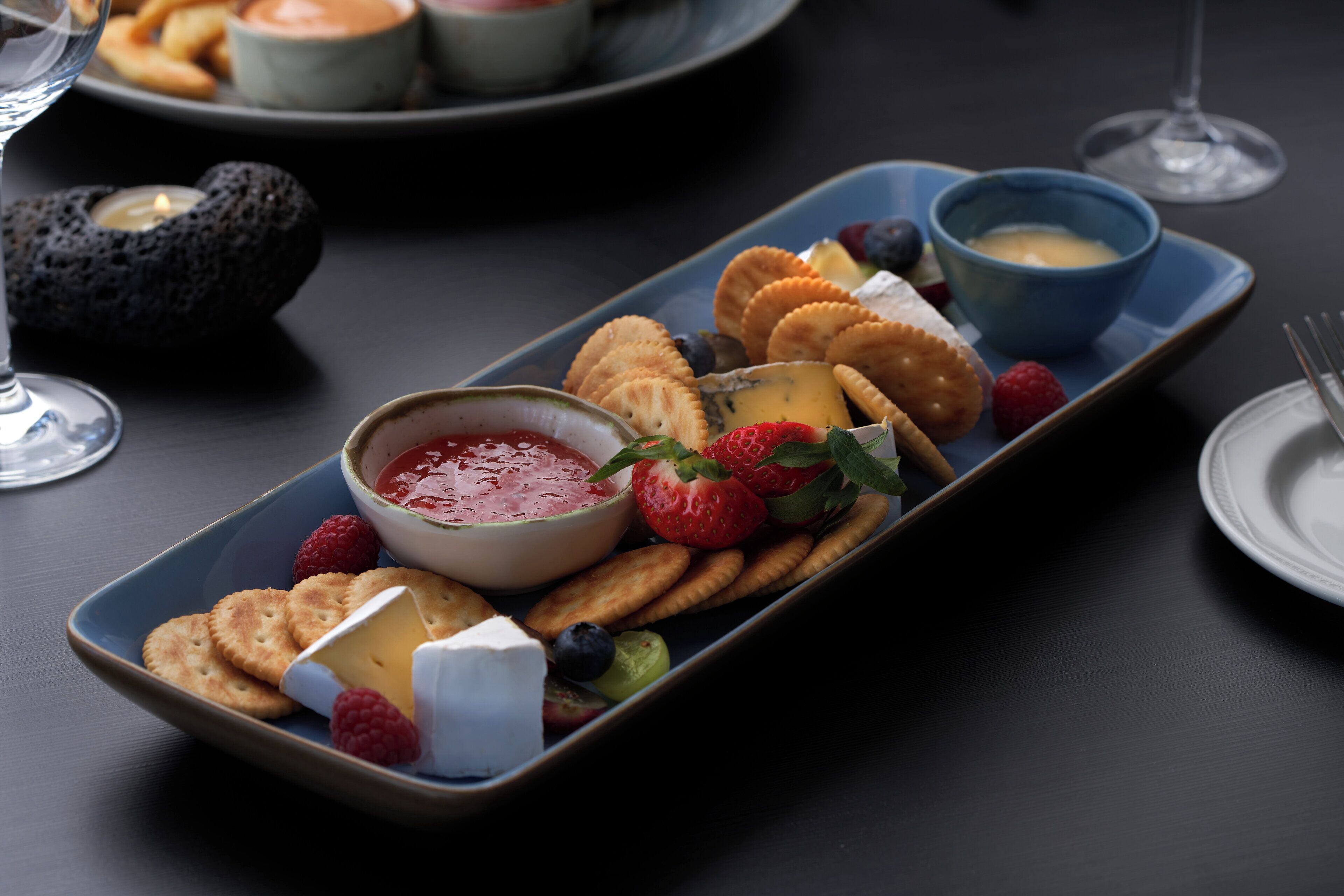 A rectangular serving tray with various cheeses, crackers, and berries, accompanied by small bowls of dips.