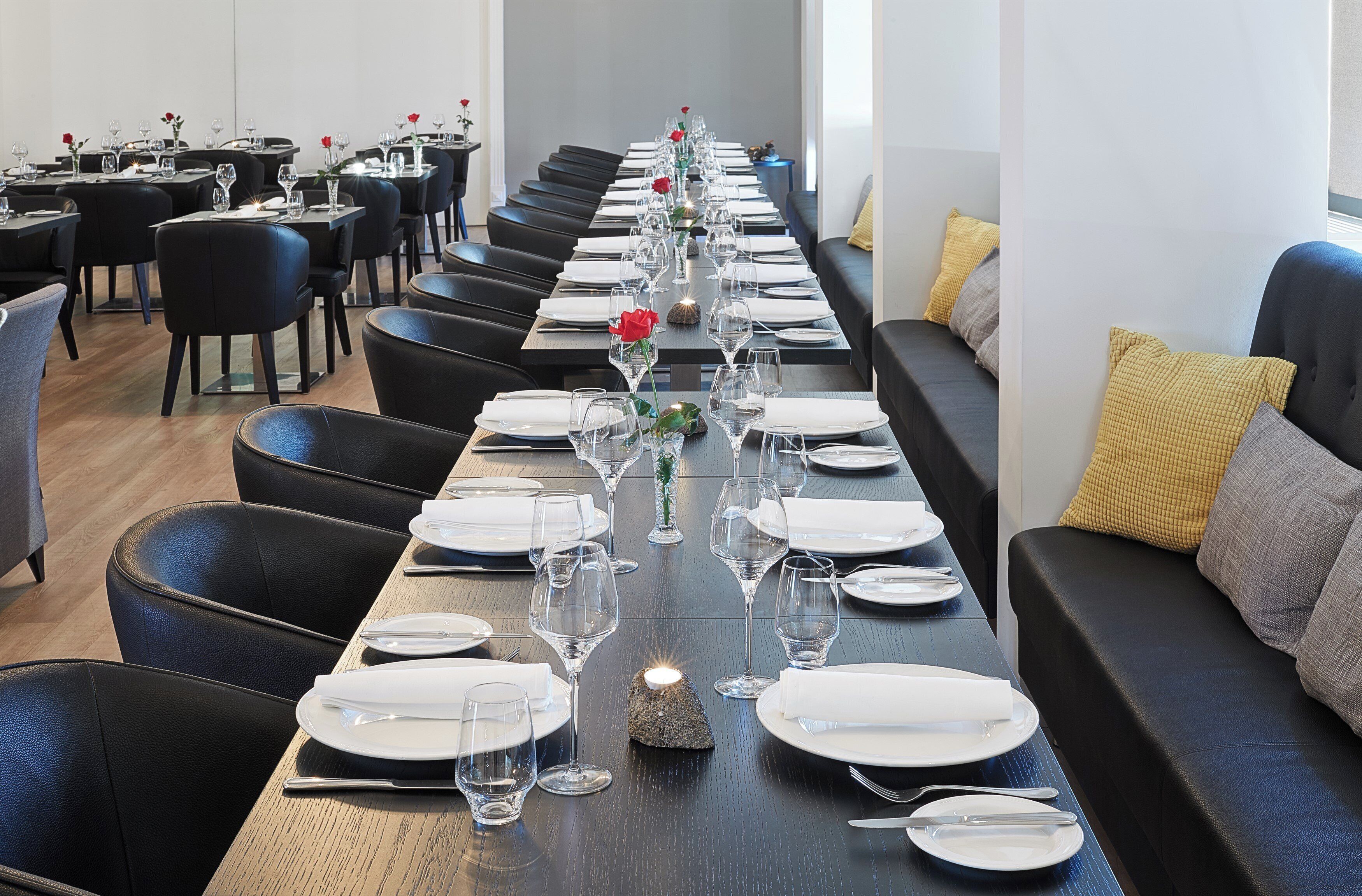 A long dining table set with white plates, glasses, and cutlery, flanked by black leather chairs and a sofa with cushions.