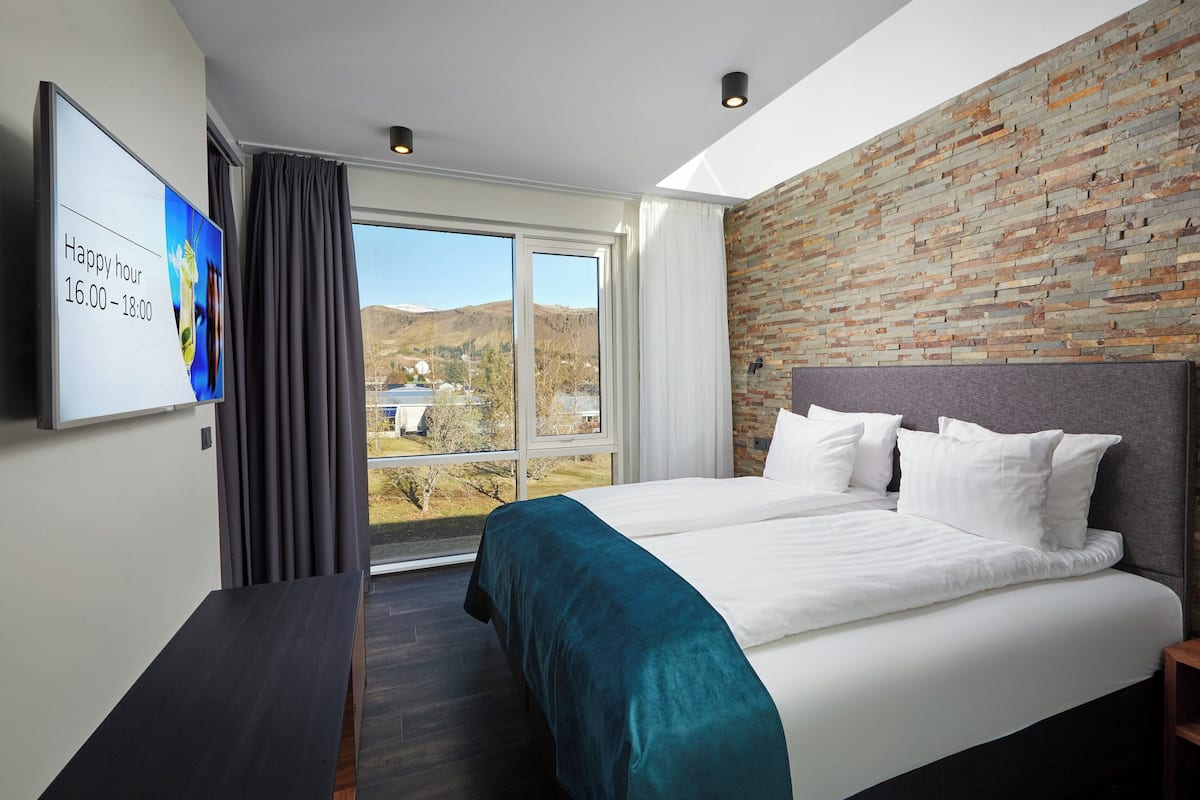 A modern hotel room with a large bed, a TV mounted on the wall, and a view of the outdoors through a large window.