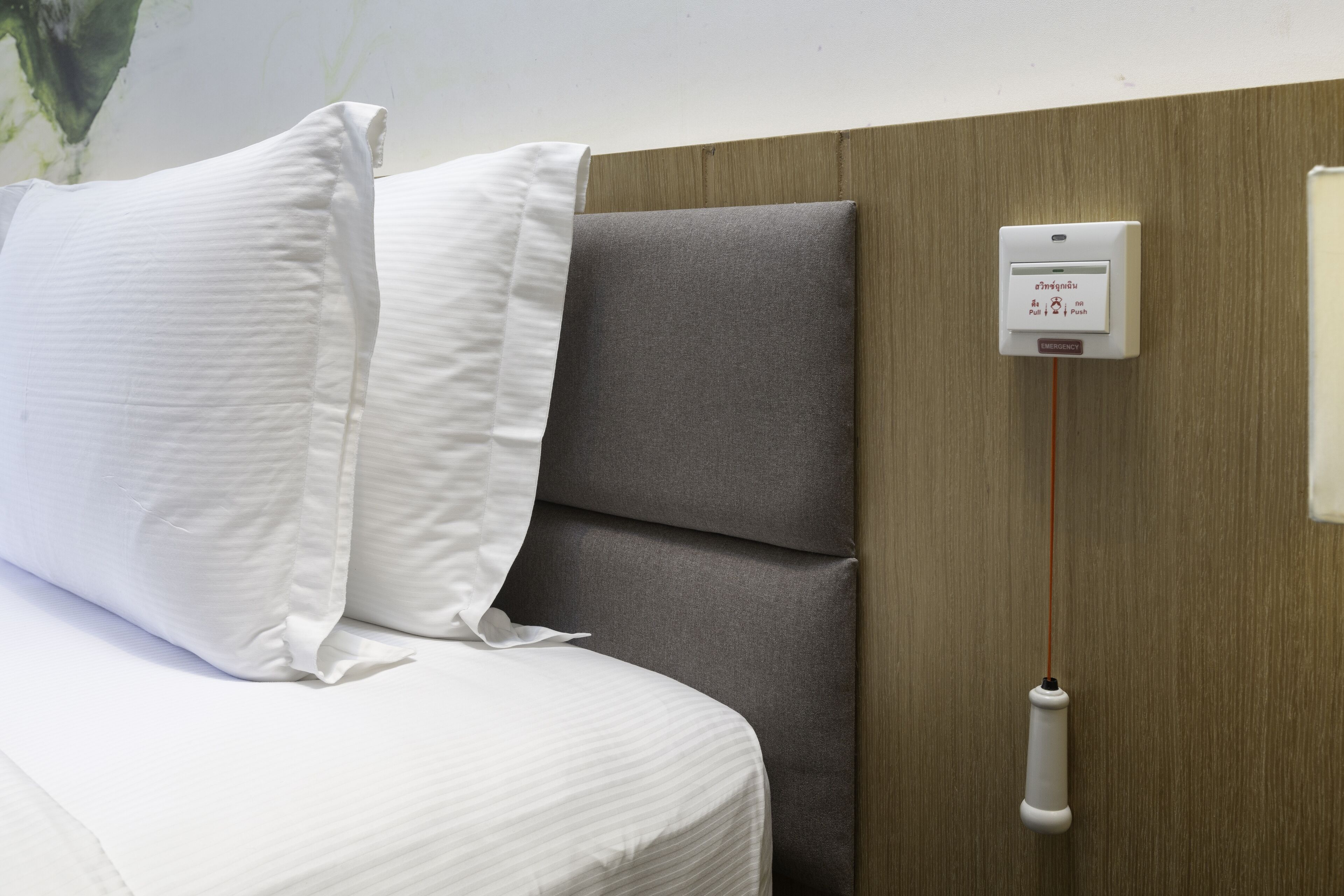 A bed with white pillows and a grey headboard, a wall-mounted emergency pull station, and a wooden panel.