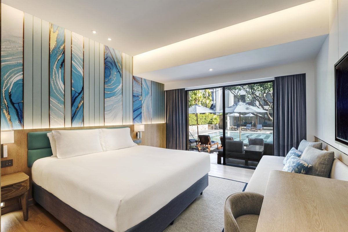 Habitación de hotel moderna con una cama grande, un sofá, un televisor y vistas al área de la piscina exterior.