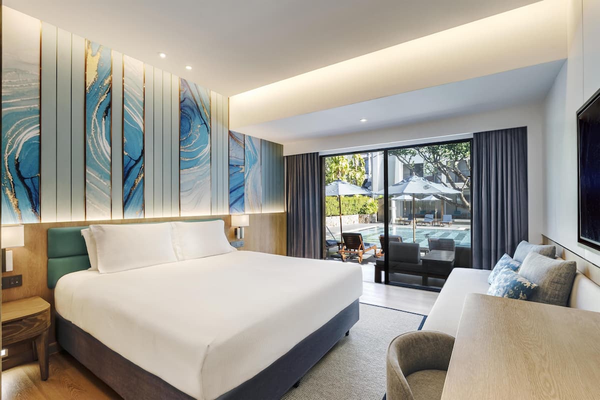 A modern hotel room with a large bed, a sofa, a TV, and a view of the outdoor pool area.