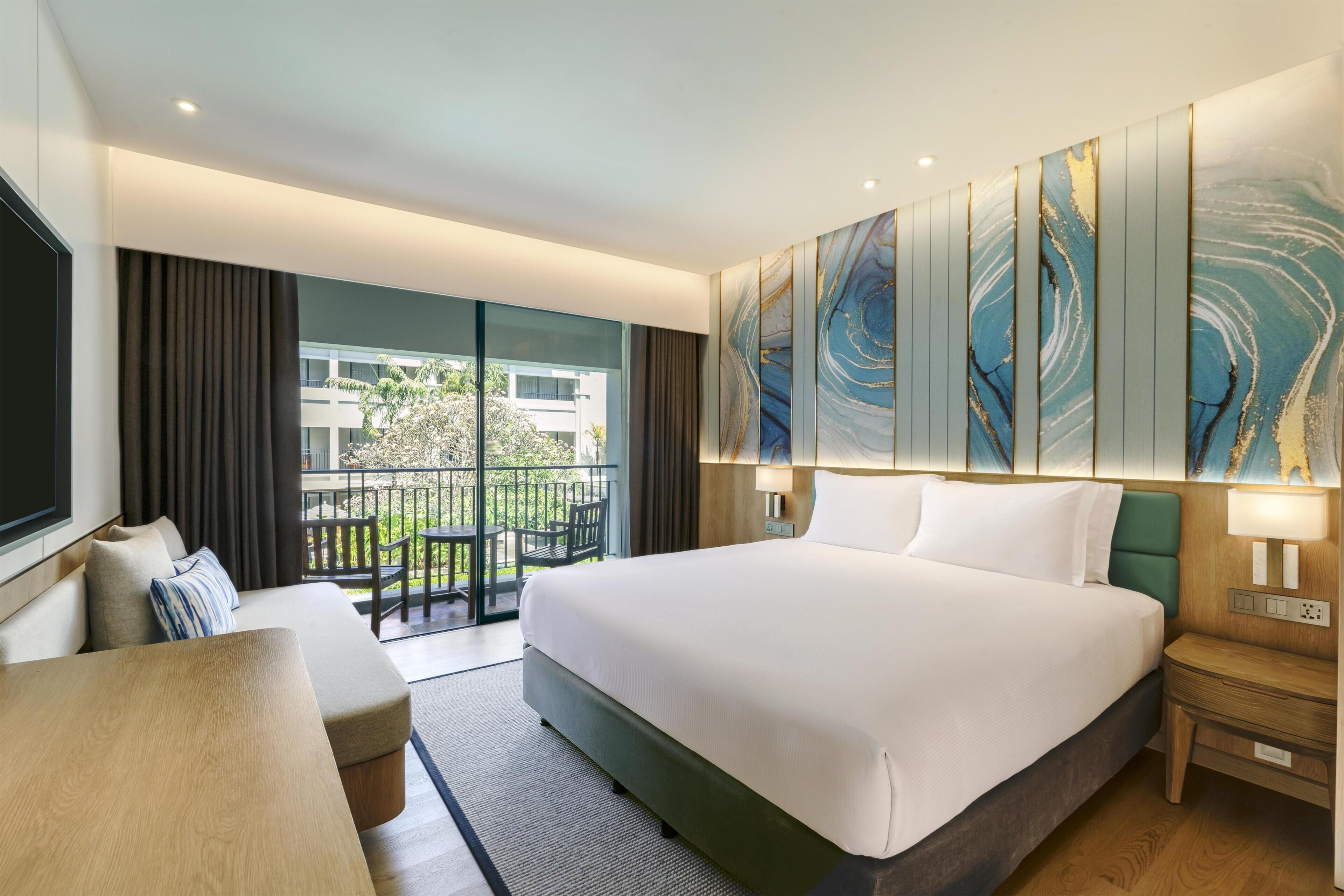 Habitación de hotel con una cama grande, un sofá, un escritorio y un balcón con vistas a edificios y zonas verdes.