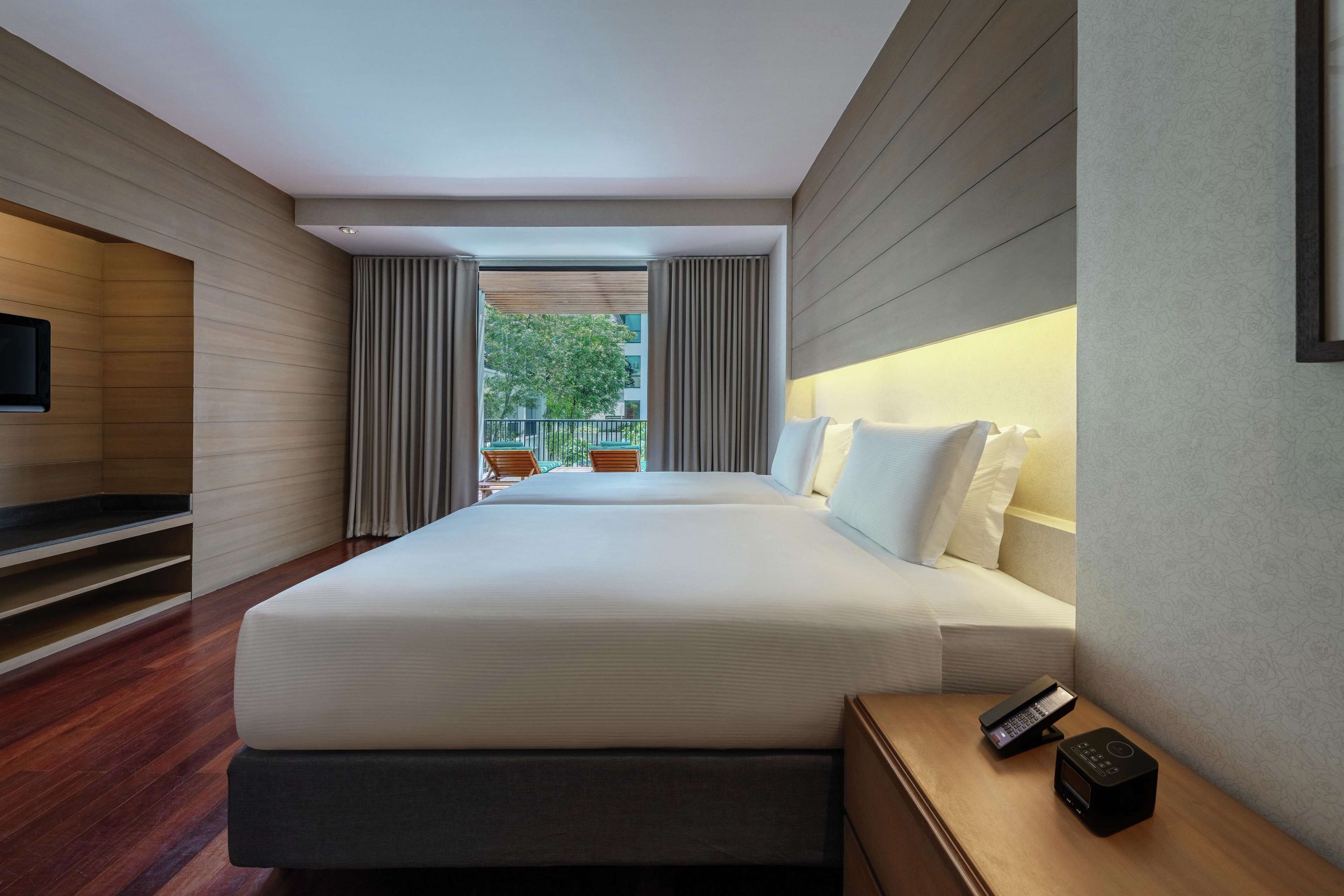 Una habitación de hotel moderna con una cama grande, una mesita de noche con un teléfono y un altavoz, un televisor montado en la pared y vistas al exterior.