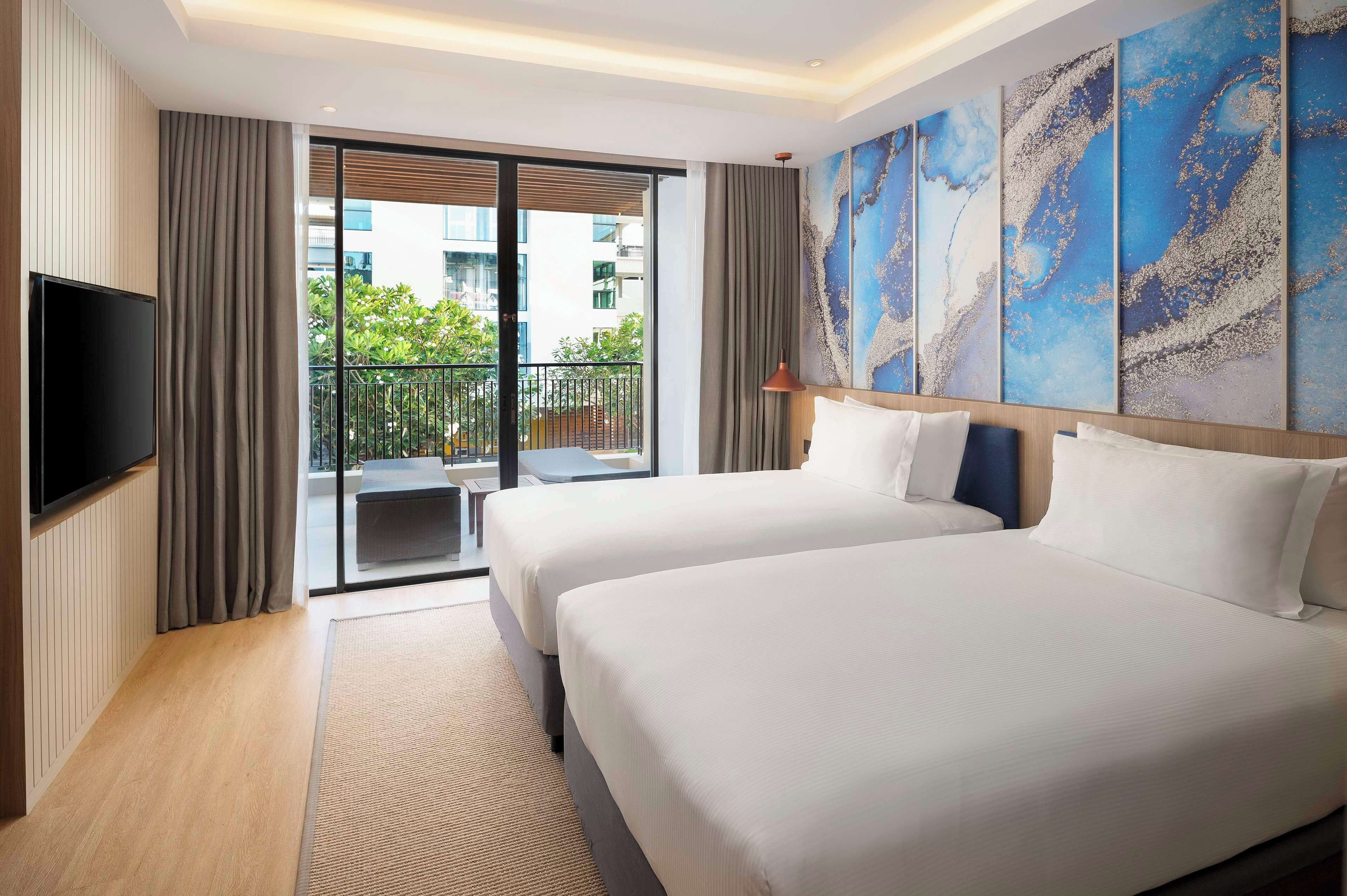 A hotel room with two beds, a flat-screen TV, a large window with a view of the city, and a balcony.