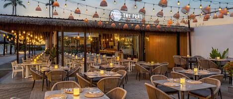 Uma área de refeições ao ar livre com mesas, cadeiras, luzes de corda e um letreiro de restaurante.