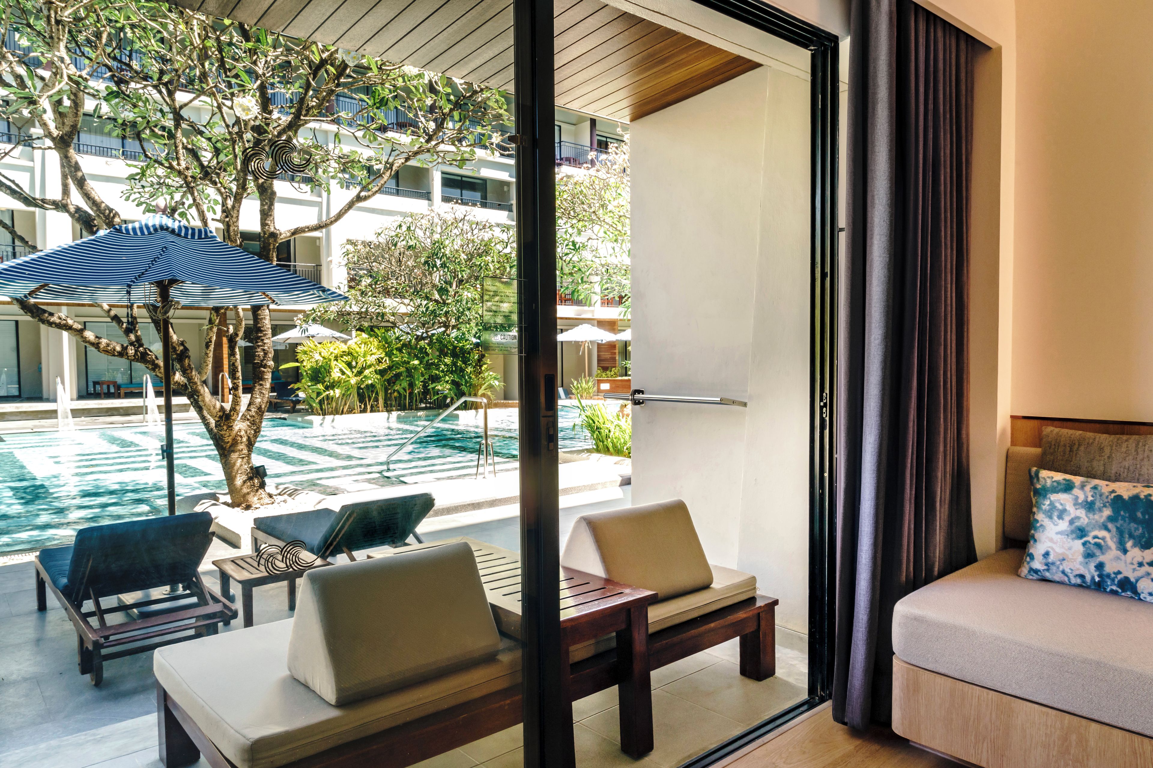 Una camera d'albergo moderna con vista su una piscina all'aperto, una zona pranzo con tavolo e sedie, e un divano con un cuscino.