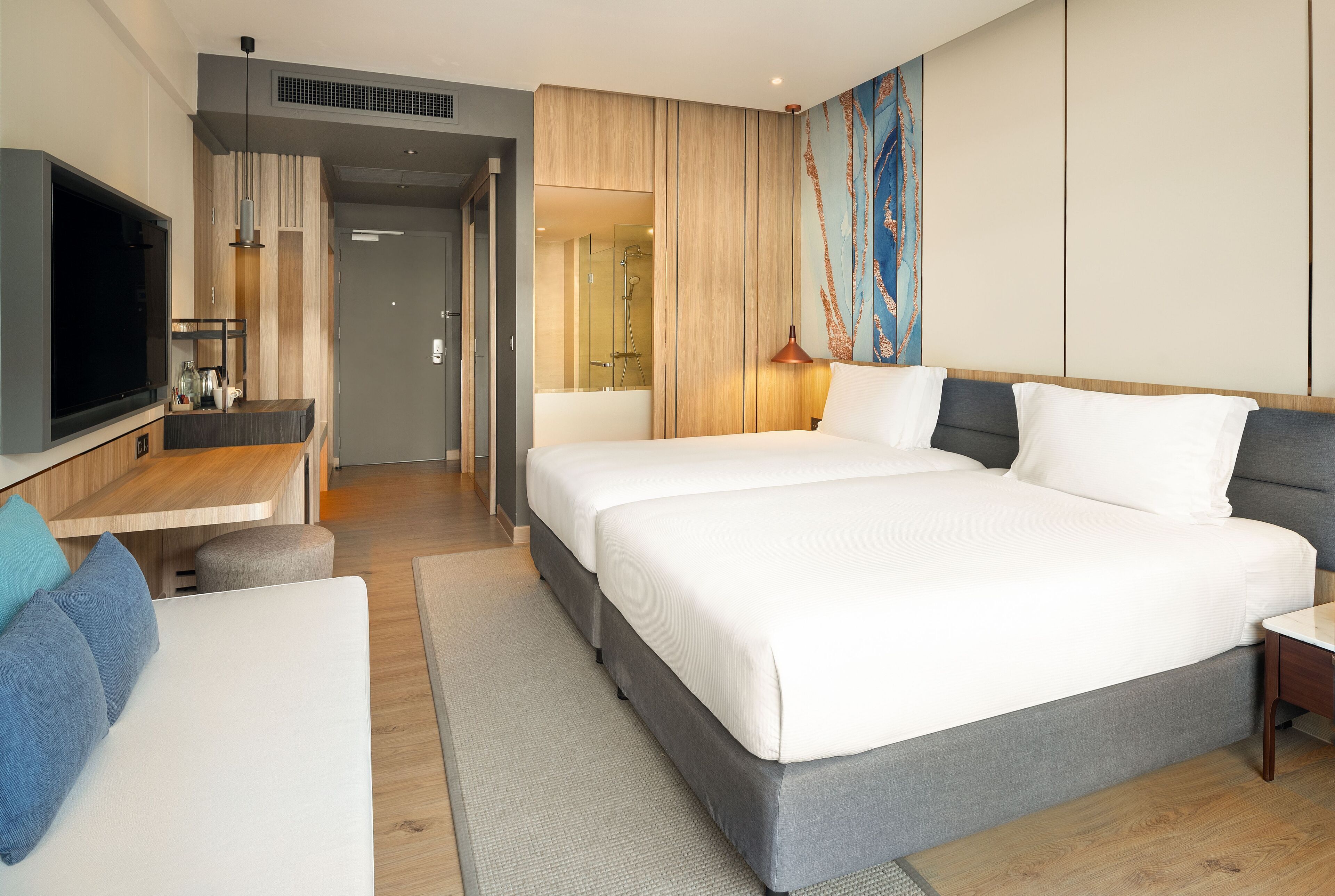 Quarto de hotel moderno com uma cama grande, uma escrivaninha com televisão de tela plana, um espaço de estar com sofá e um banheiro visível através de uma porta aberta.