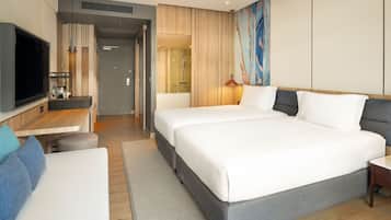Quarto de hotel moderno com uma cama grande, uma escrivaninha com televisão de tela plana, um espaço de estar com sofá e um banheiro visível através de uma porta aberta.