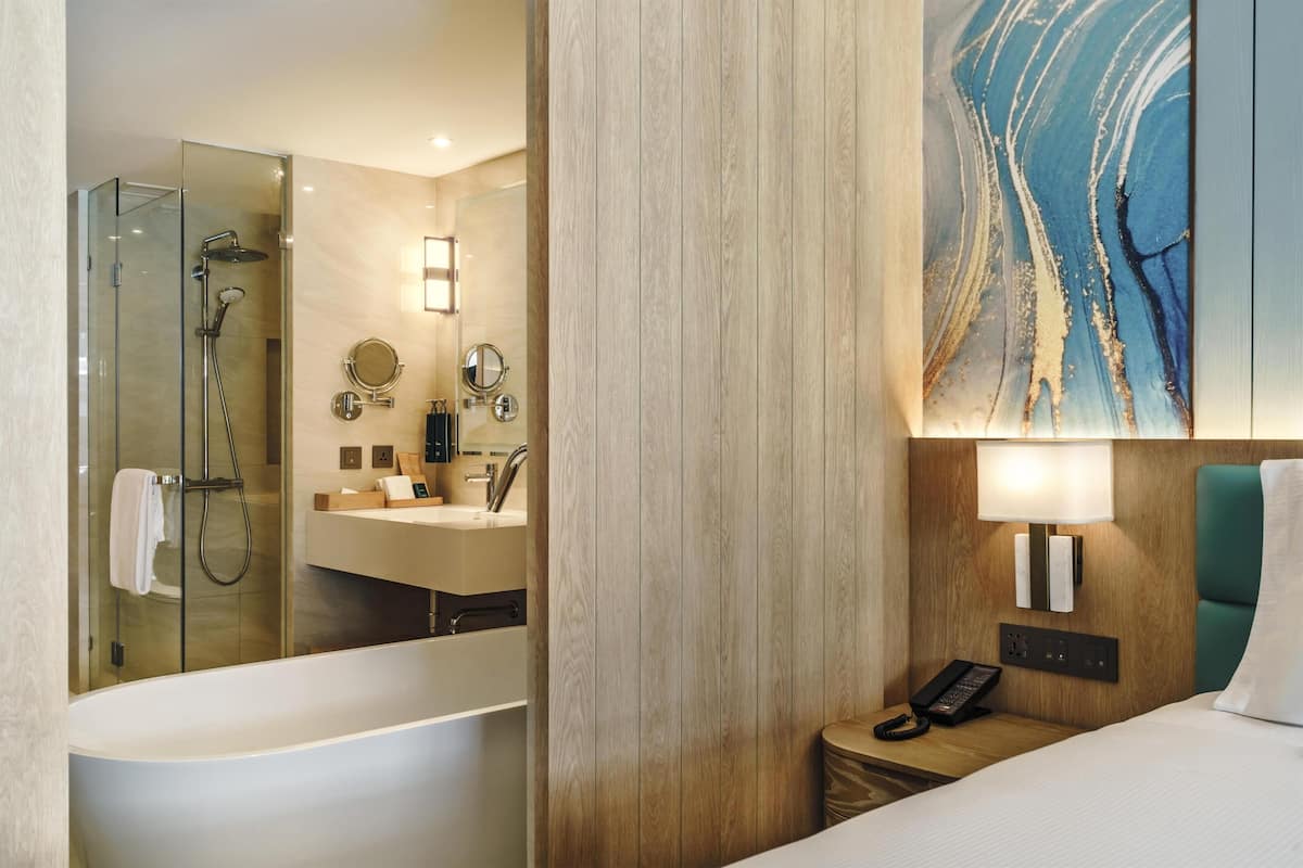 Habitación de hotel moderna con ducha de vidrio, una bañera grande, panel de pared de madera, un cuadro, una lámpara de noche y una mesita con un teléfono.