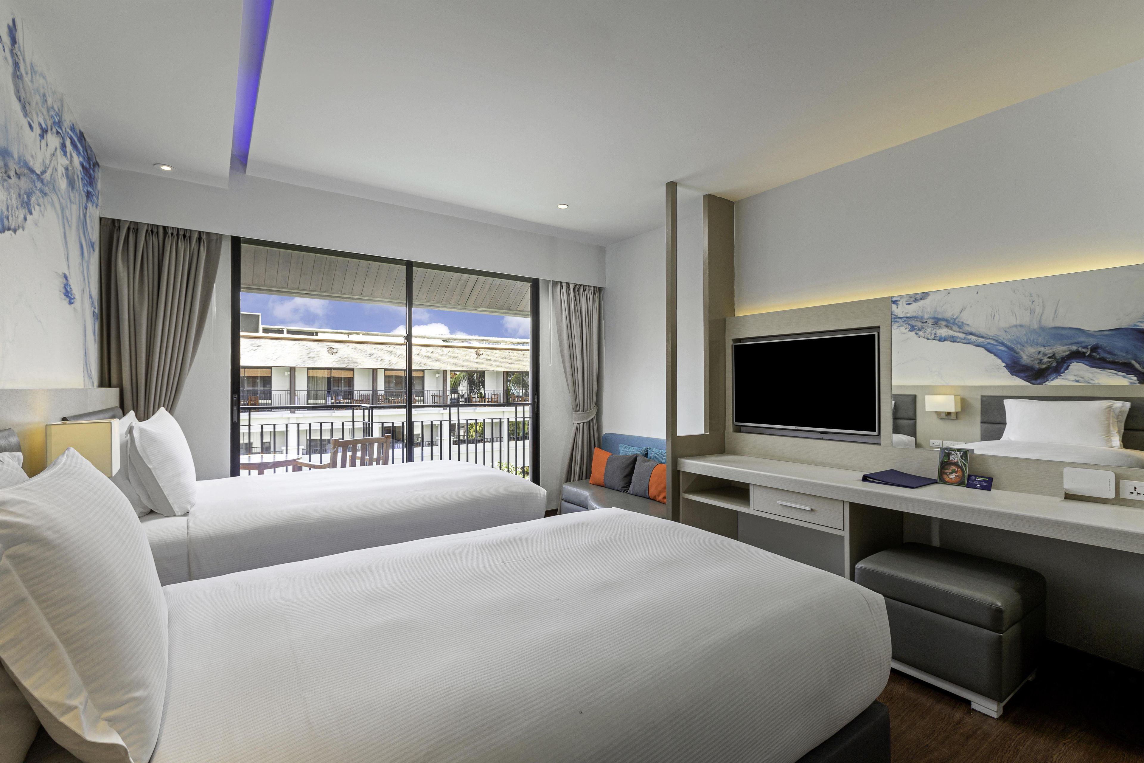 A hotel room with two beds, a desk, a flat-screen TV, and a view of the building's exterior.